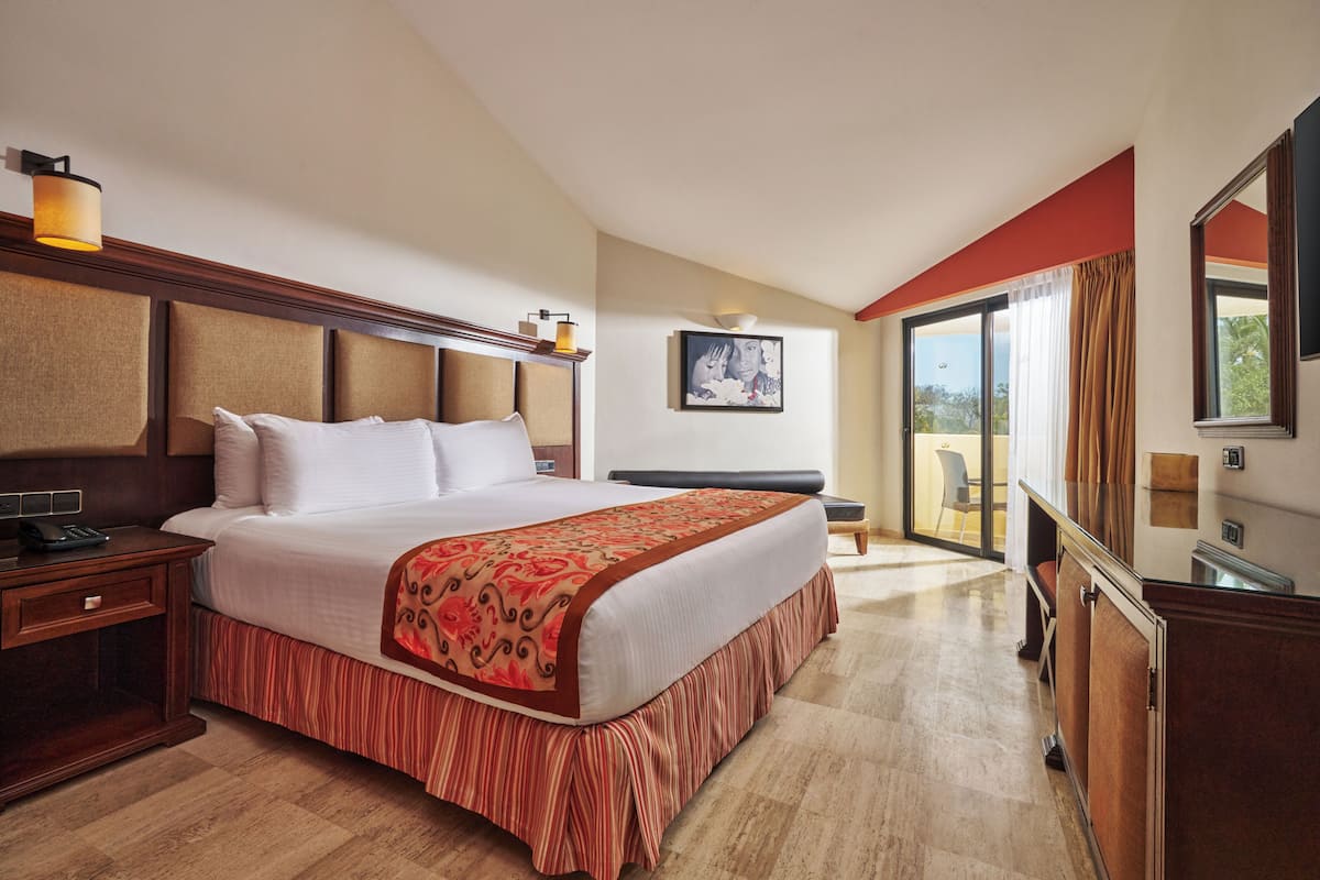 A hotel room with a large bed, a bedside table with a lamp, a chair, and a large window with curtains.