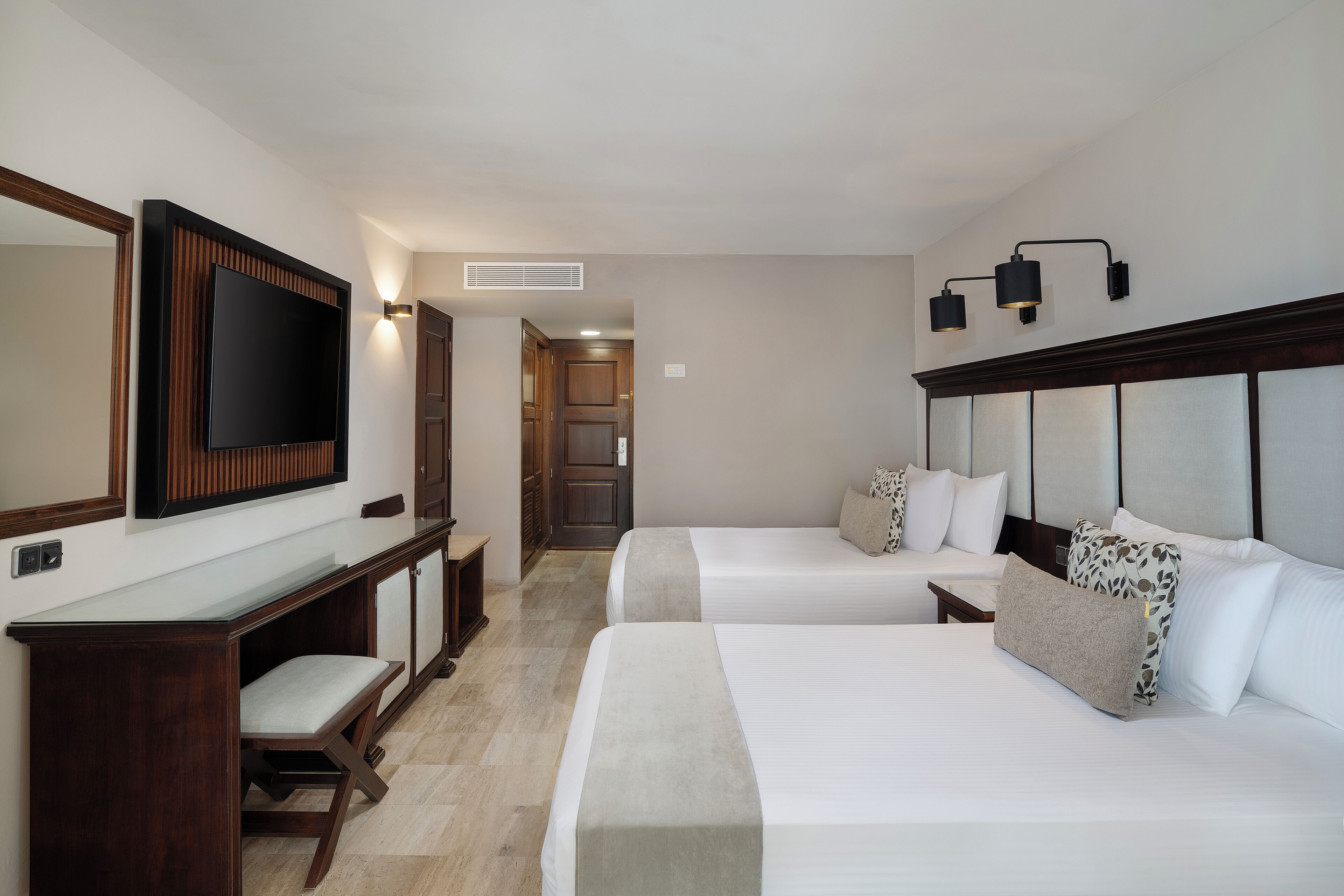 A hotel room with two beds, a desk, a chair, a large mirror, and a television.
