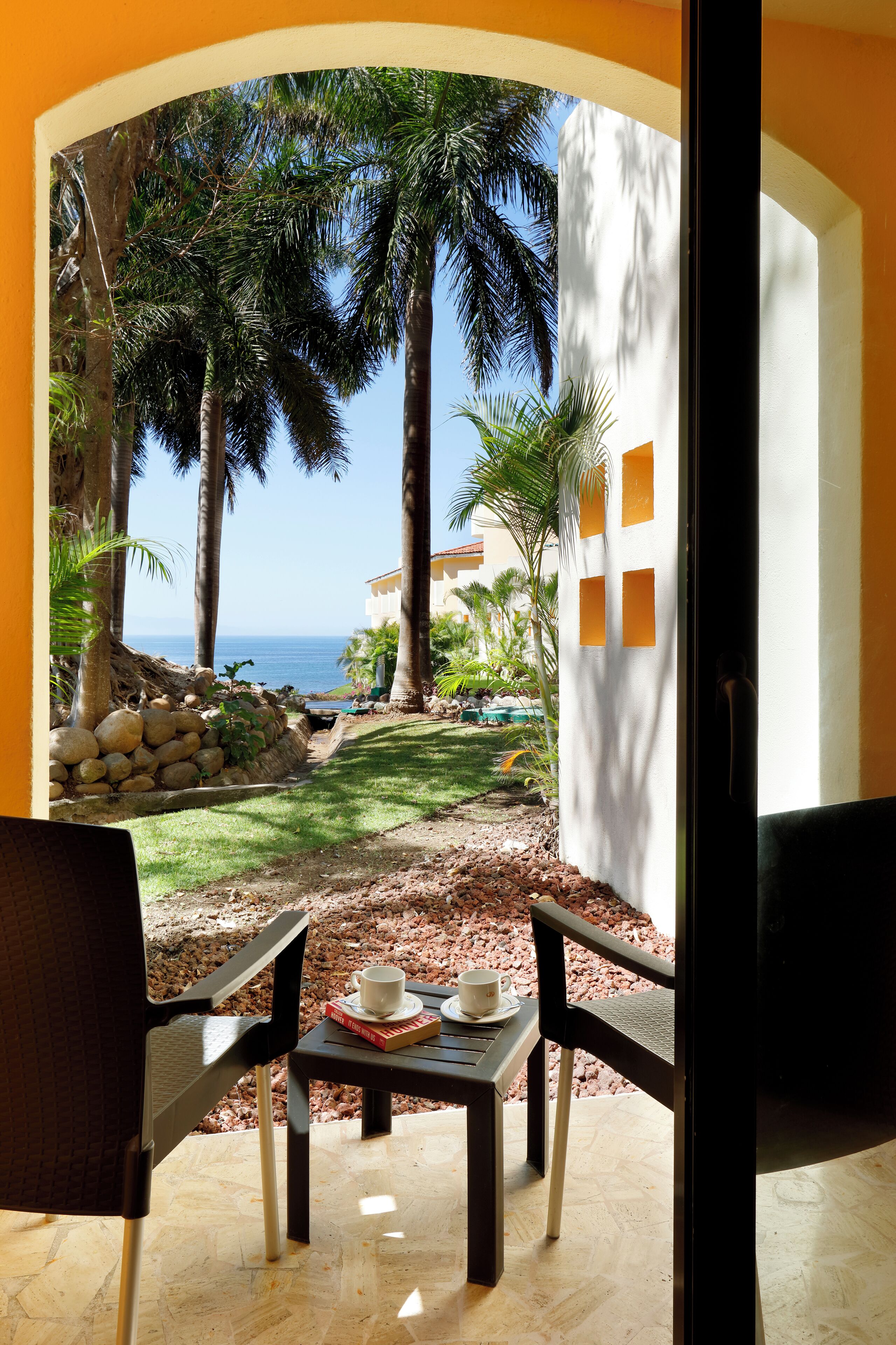 A patio with two chairs and a table, overlooking a garden with palm trees and a view of the ocean.