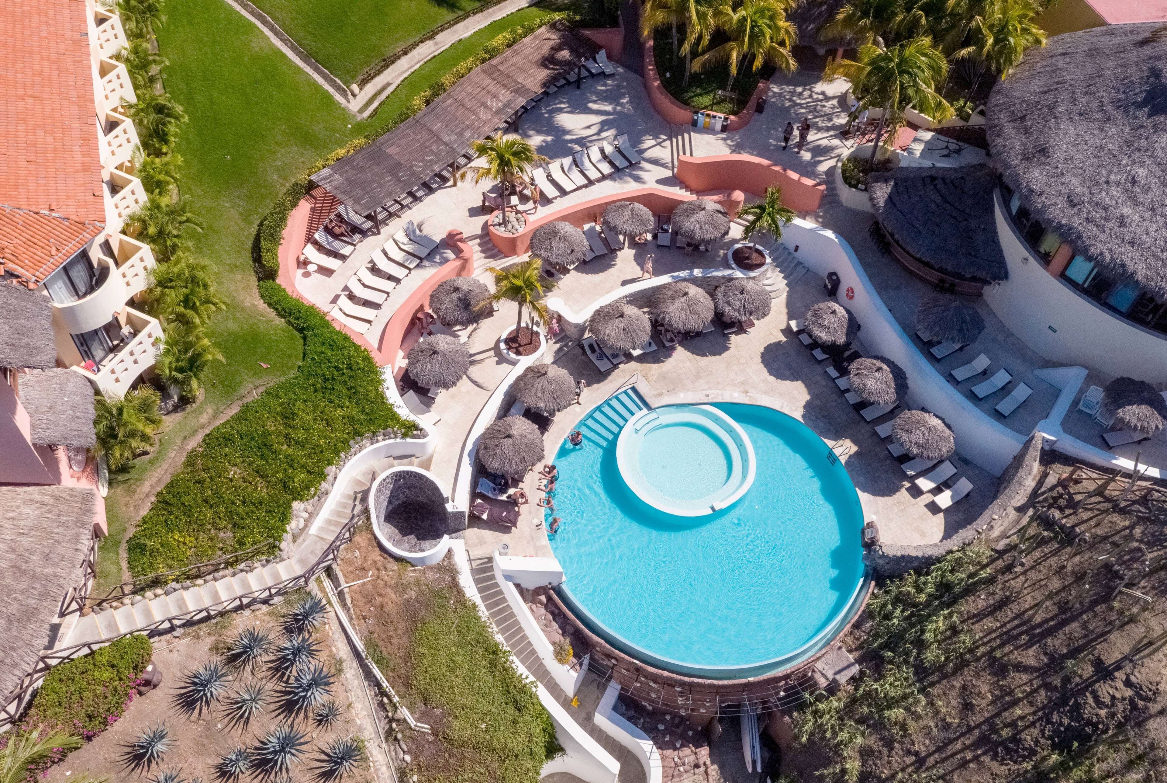 Vista aérea de un resort con una piscina central, áreas de descanso con sombrillas de paja y palmeras.