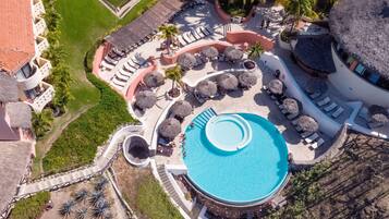 Vista aérea de un resort con una piscina central, áreas de descanso con sombrillas de paja y palmeras.