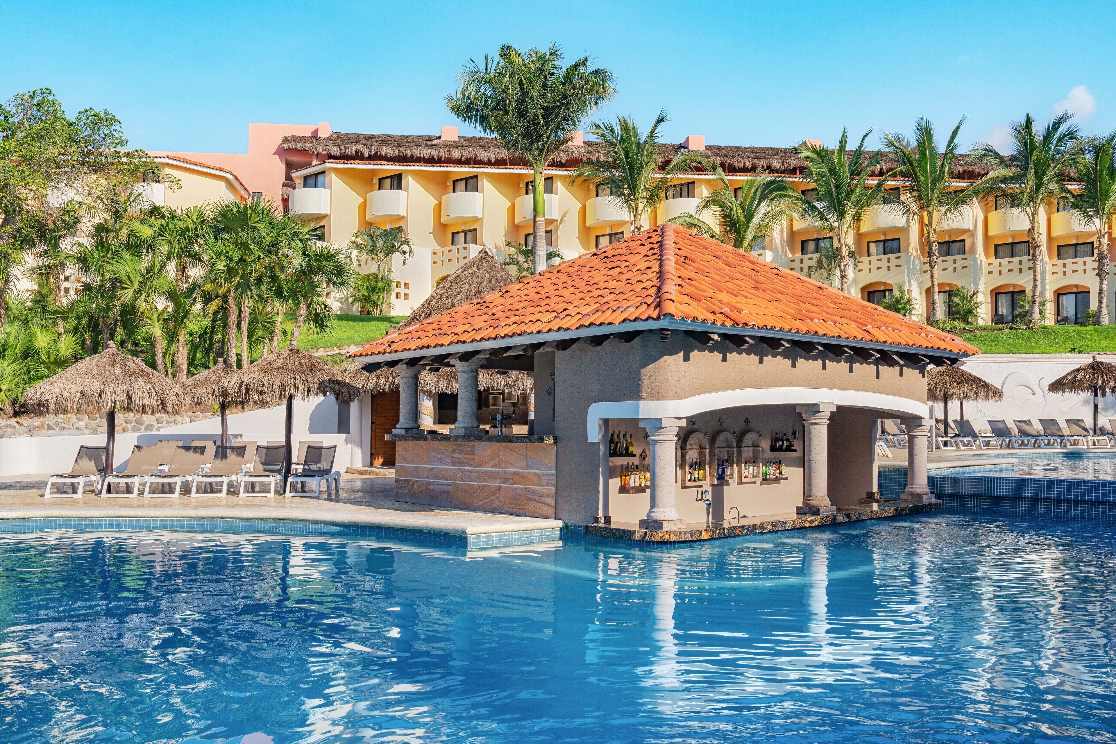 A pool area with lounge chairs, a bar, and a building with a tiled roof.