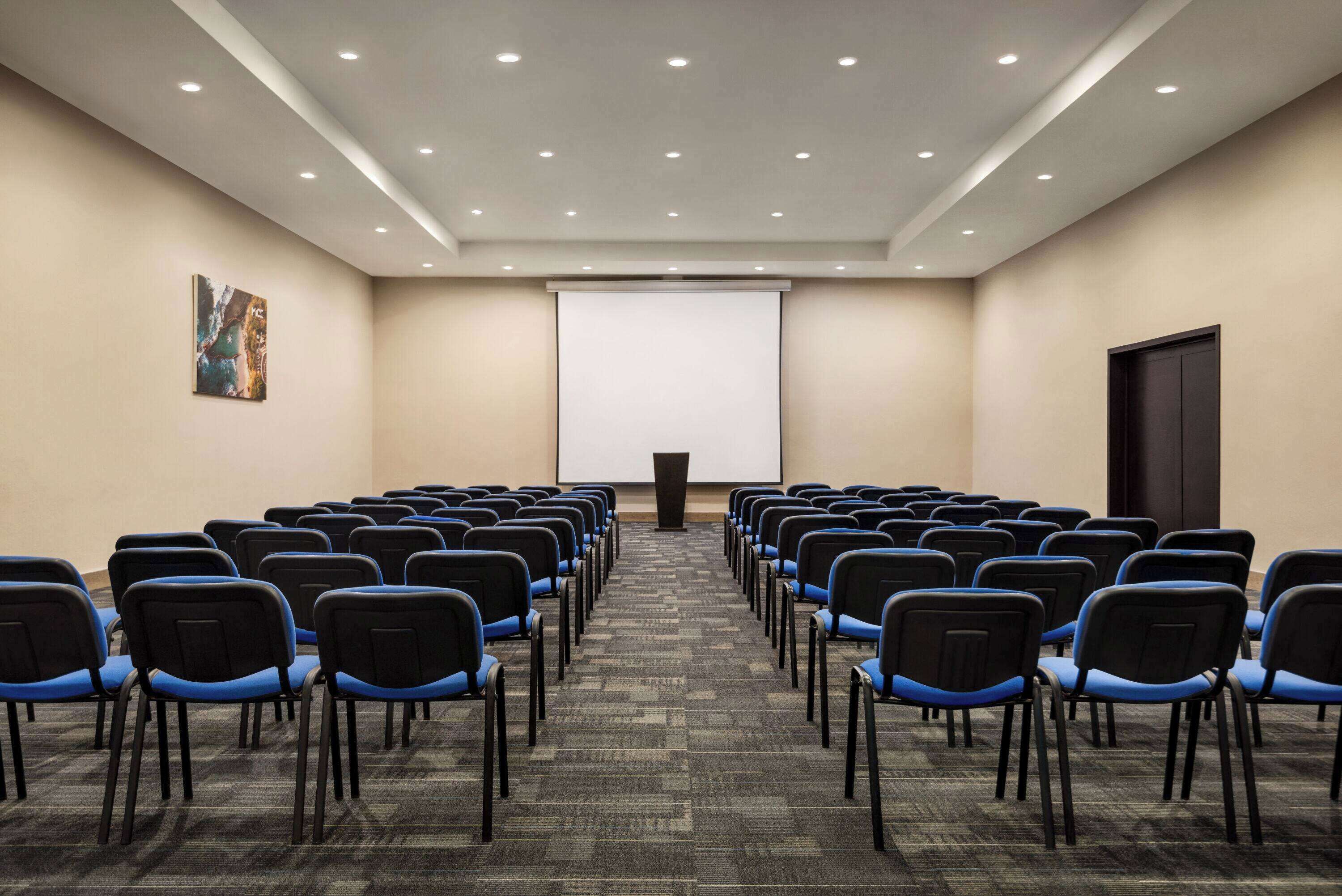 Een congrescentrum met rijen blauwe stoelen, een scherm voor een beamer en een vloerbedekking.