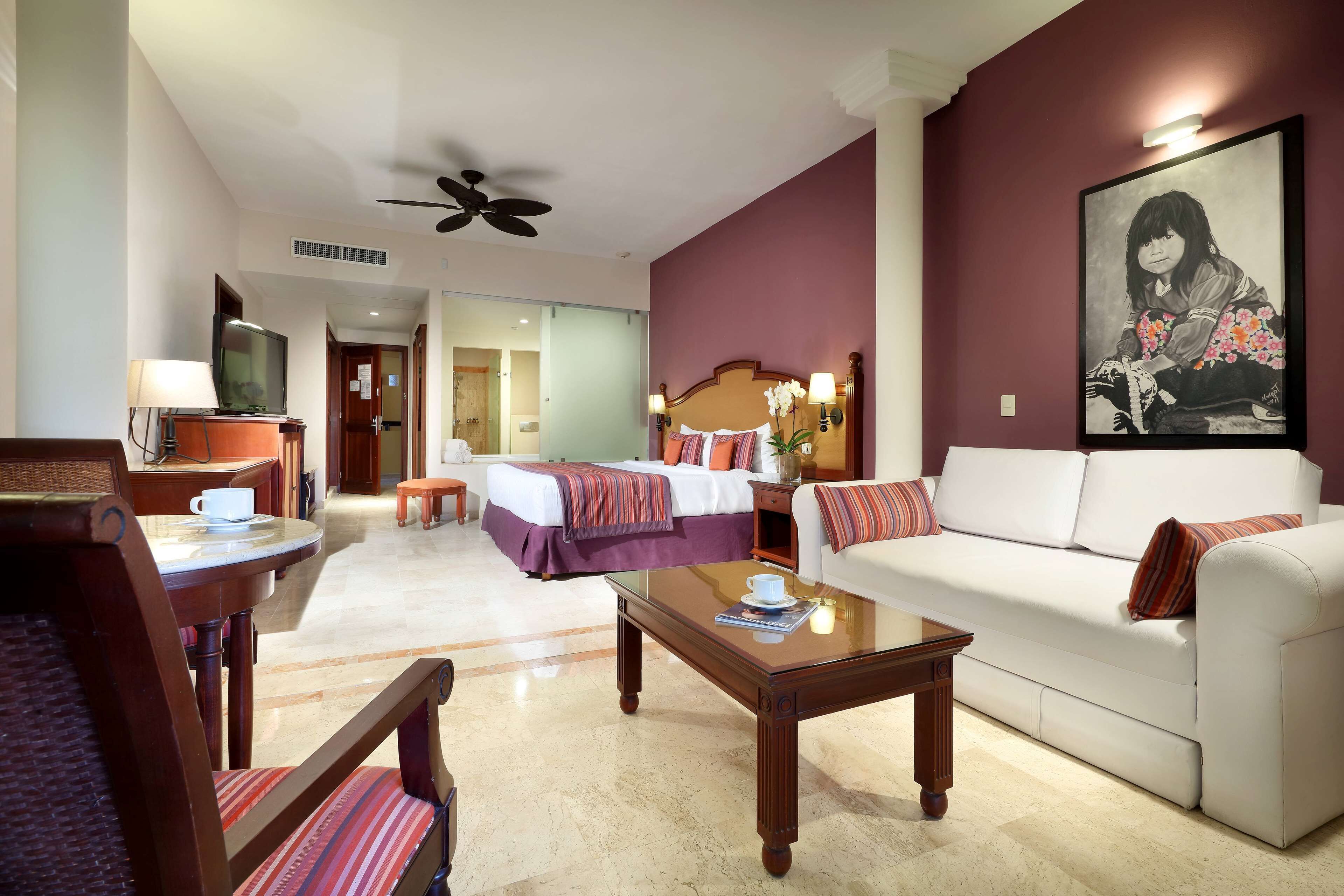 A hotel room with a bed, a sofa, a coffee table, a TV, and a painting on the wall.