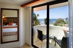 Quarto de hotel com uma cama, uma cadeira e uma mesa perto de uma porta de vidro que desliza, com vista para uma praia.