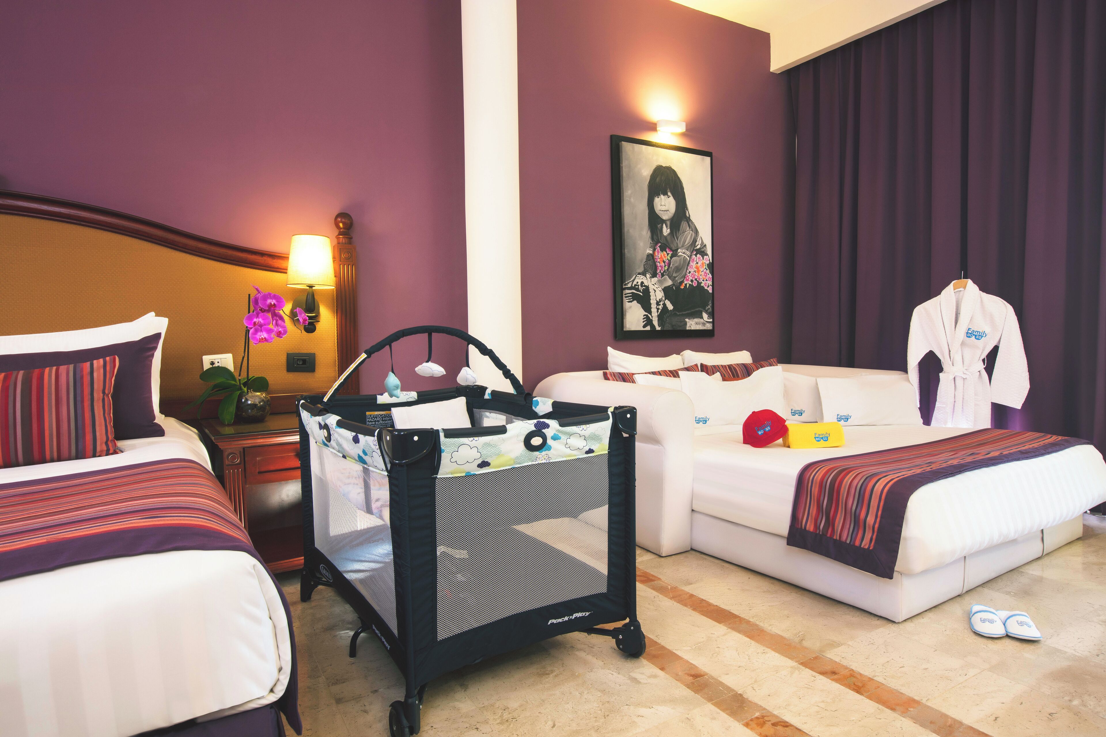 Habitación de hotel con cama, sofá, corralito para bebé, lámpara, cuadro enmarcado y abrigo en la silla.
