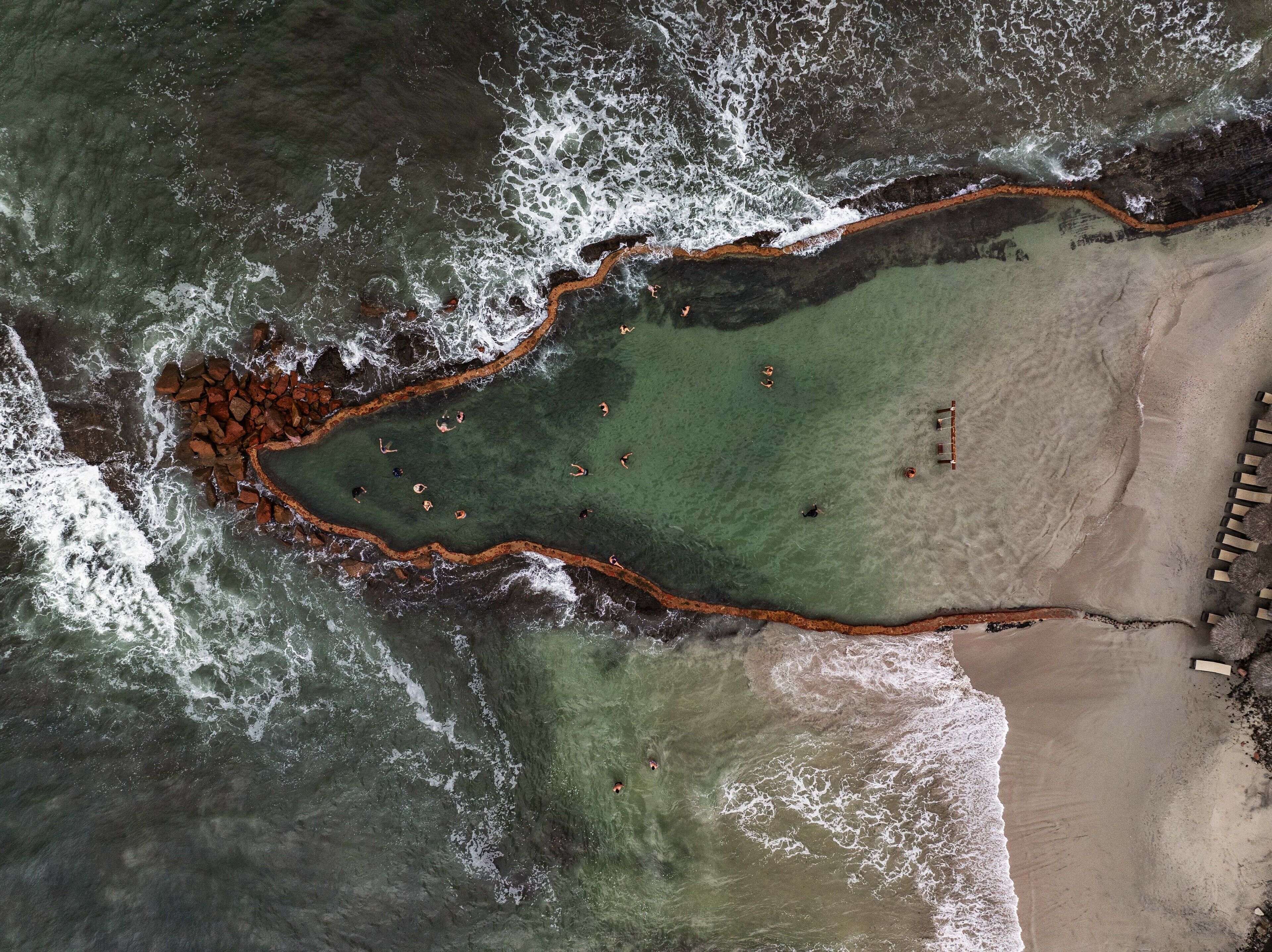 An aerial view of a beach with clear water, a rocky shoreline, and a few people swimming.