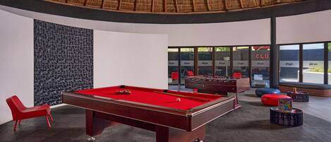 A pool table in a modern room with a geometric wall pattern, a red chair, and a large window with 'CLUB' signage.