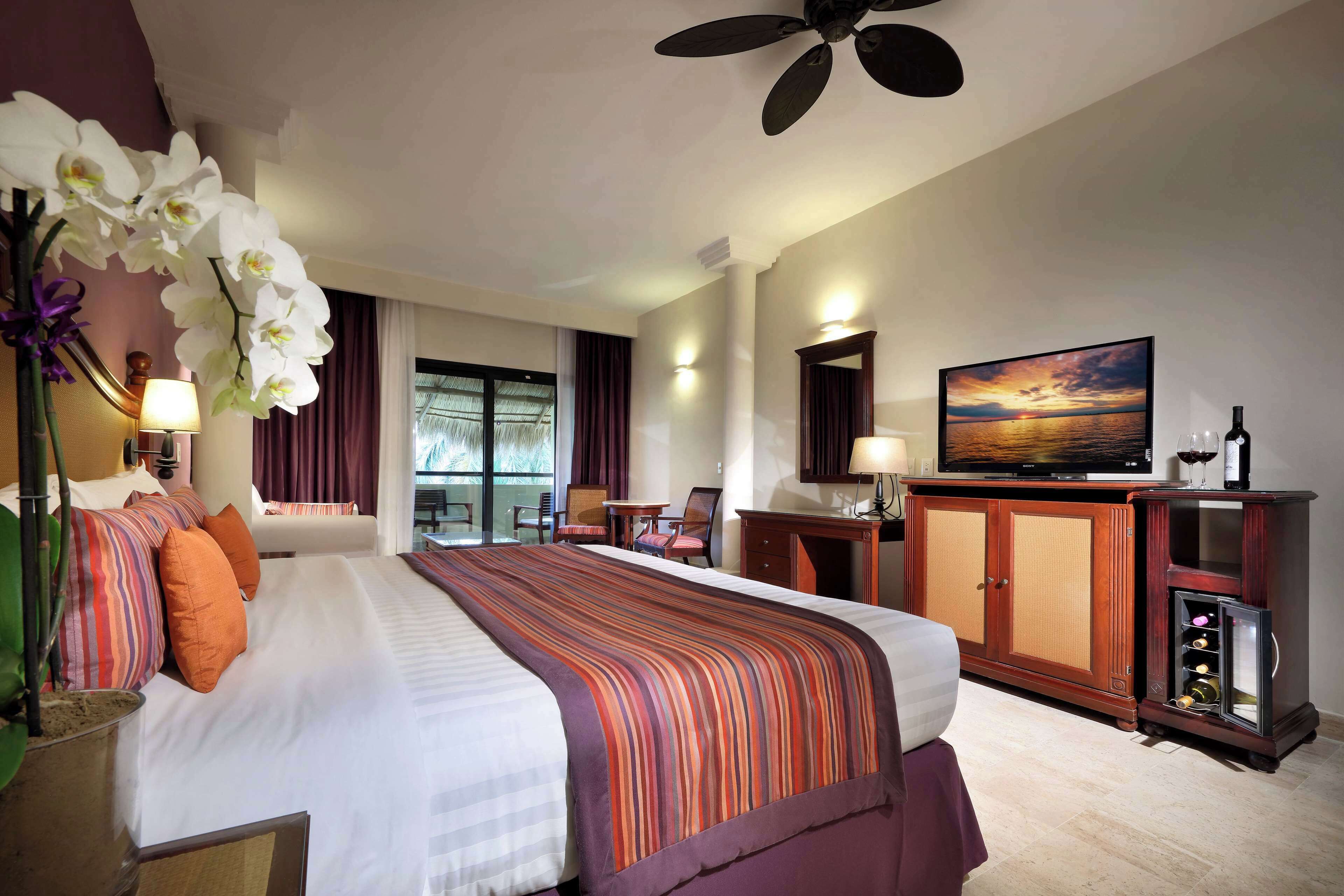 A hotel room with a large bed, a desk, a chair, a cabinet with wine bottles, and a painting on the wall.