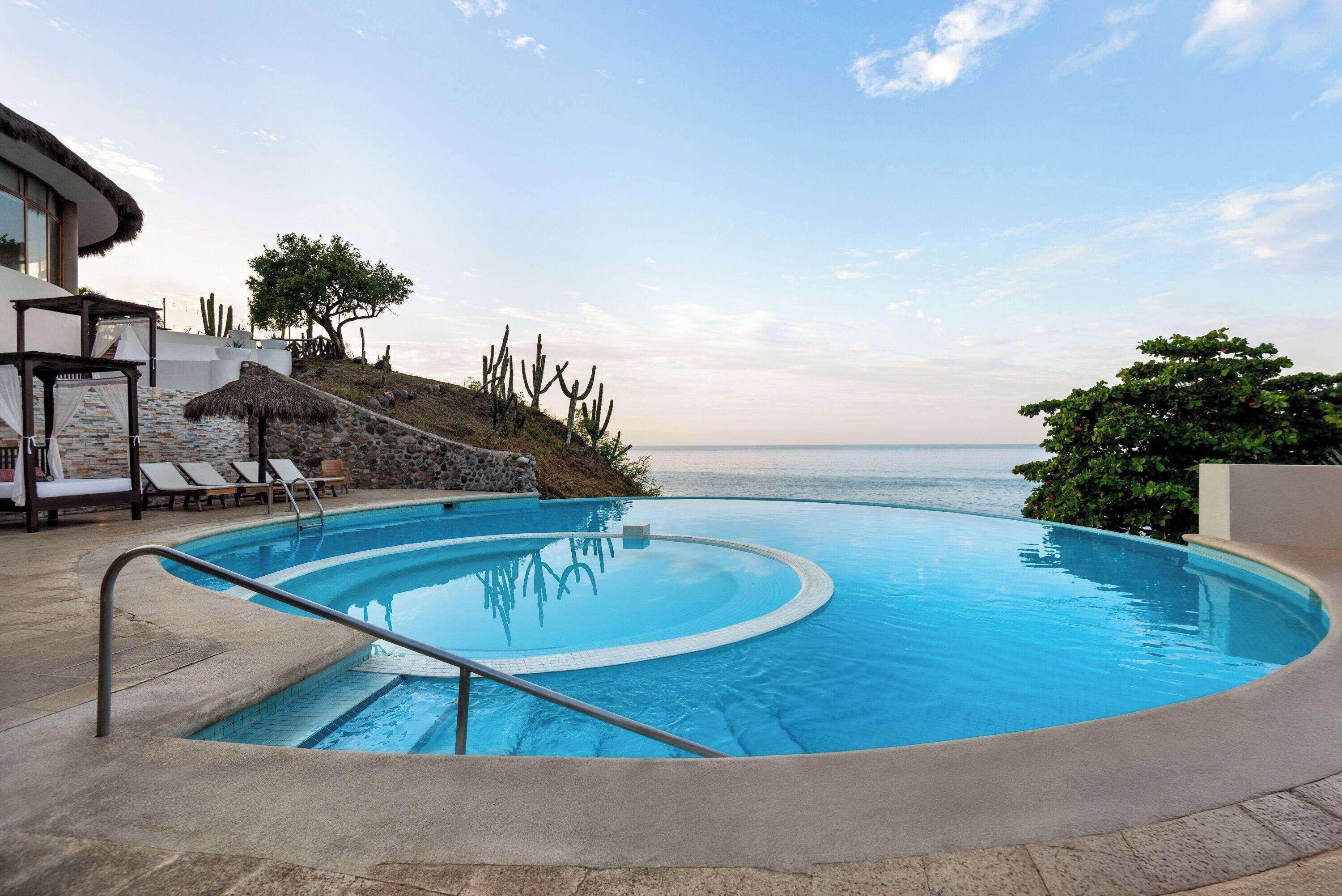 A swimming pool with a clear view of the ocean, surrounded by lounge chairs and a thatched roof structure.