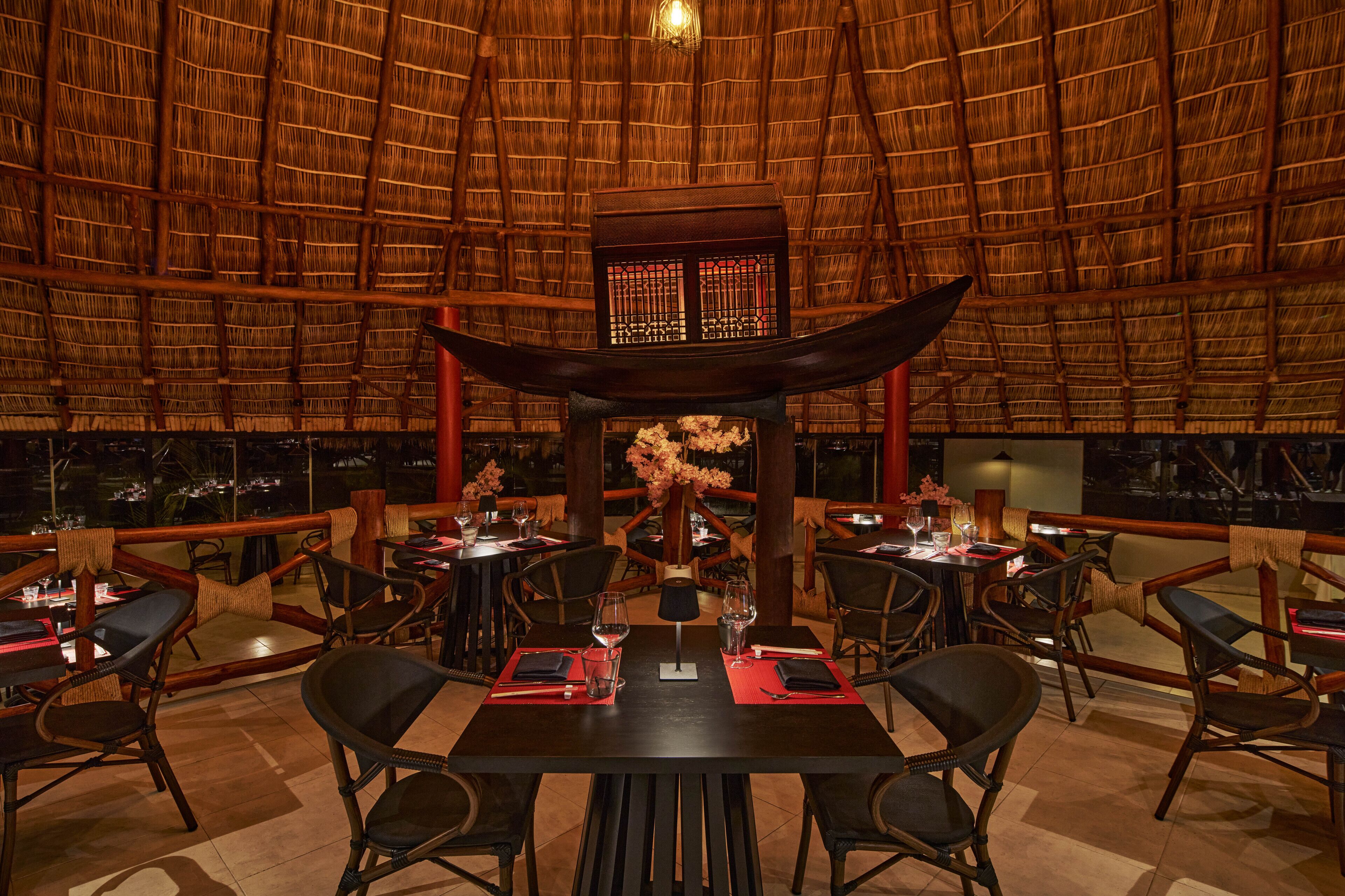 Un restaurant doté d’un toit de chaume, de meubles en bois et d’un élément décoratif central. Les tables sont dressées avec des sets de table rouges, des verres à vin et des couverts.