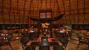 Un restaurant doté d’un toit de chaume, de meubles en bois et d’un élément décoratif central. Les tables sont dressées avec des sets de table rouges, des verres à vin et des couverts.