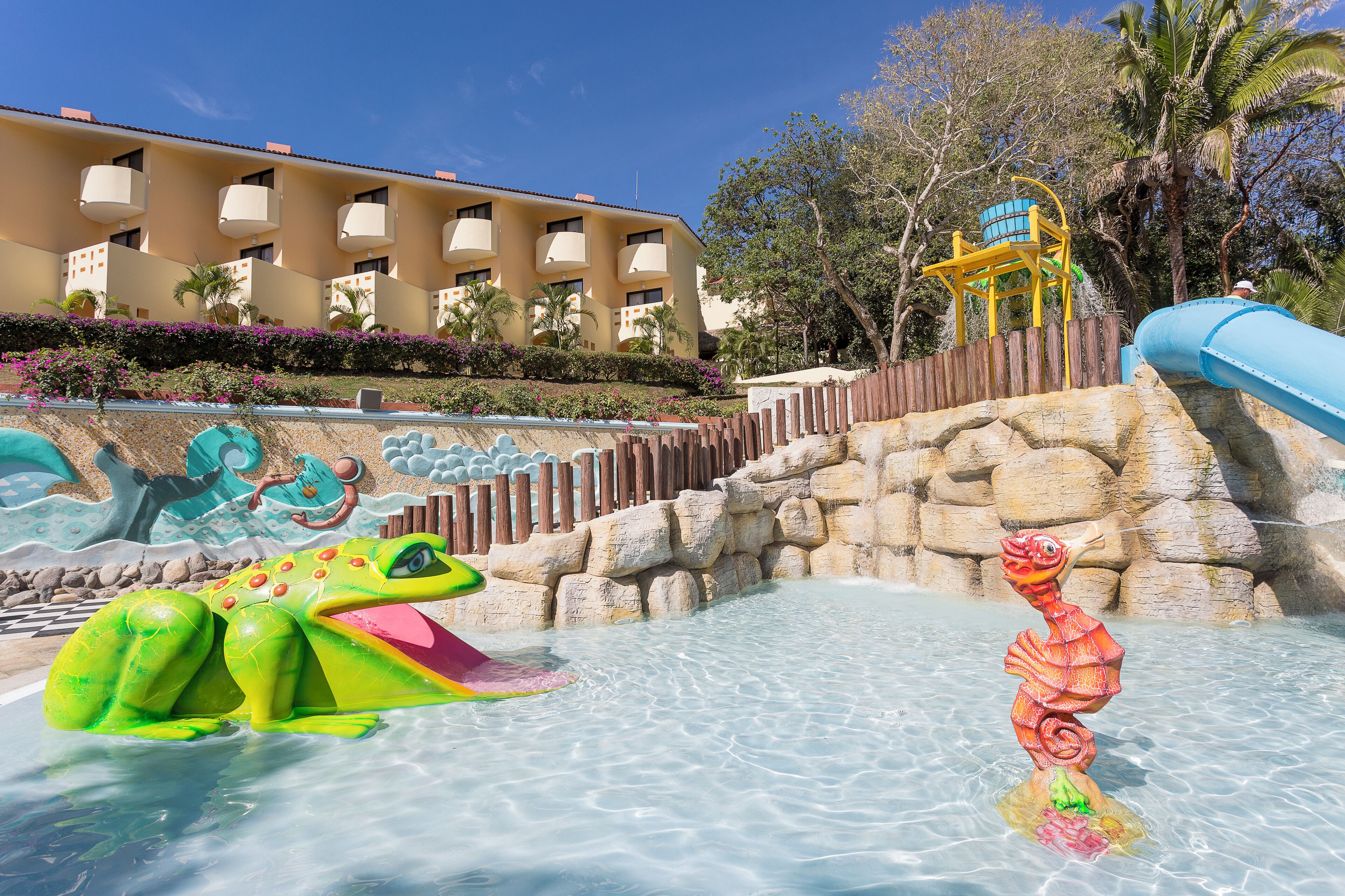 A water park with a large building in the background, a slide, and a frog-shaped water feature.
