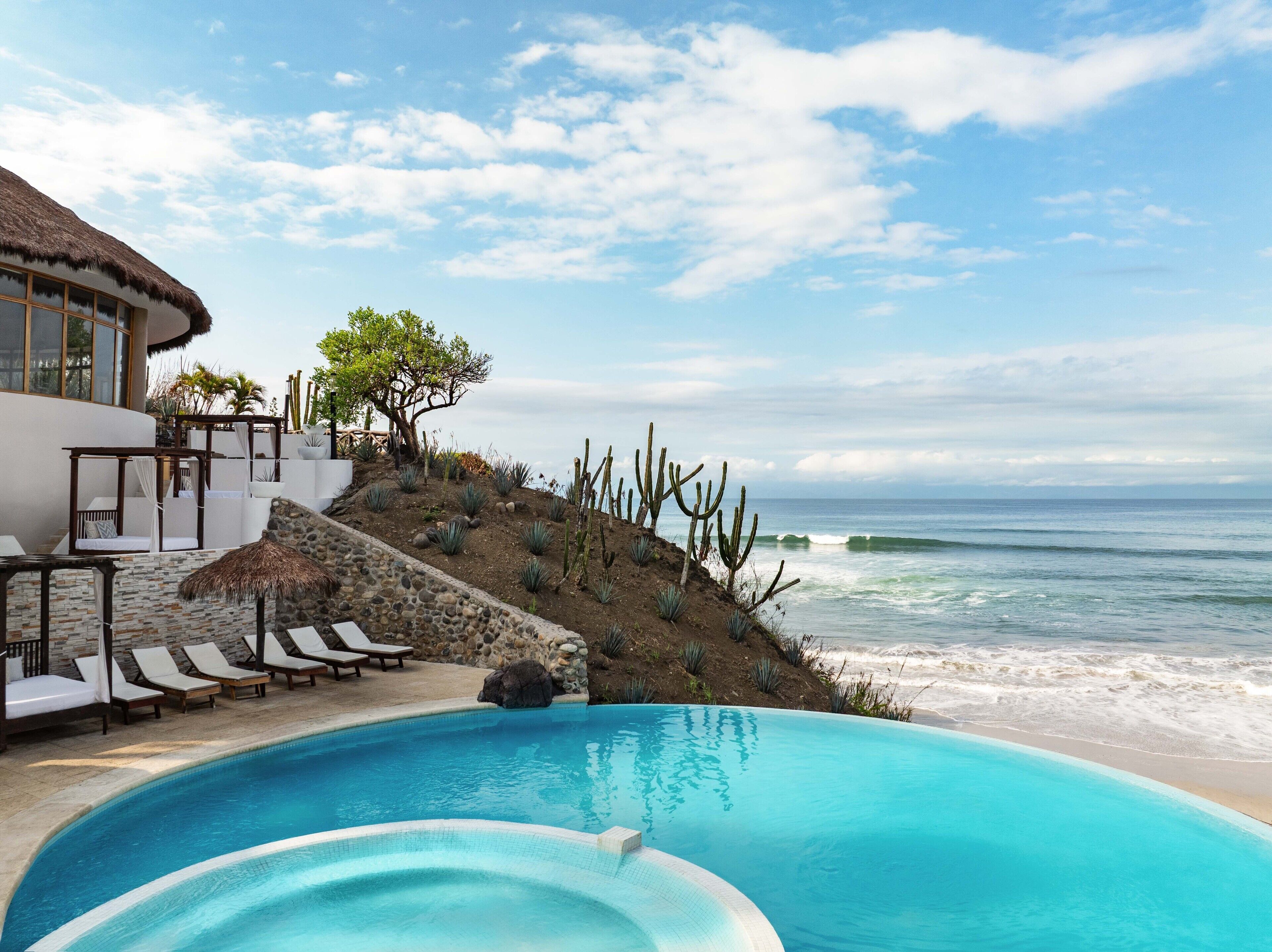 Une piscine avec vue sur l’océan, une terrasse avec des chaises longues et un bâtiment doté d’un toit de chaume.