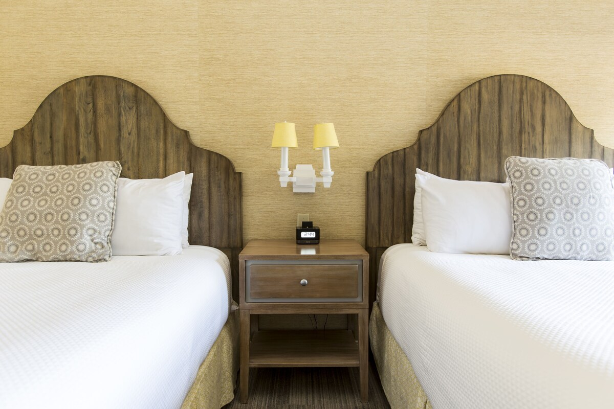Habitación de hotel con dos camas, cada una con almohadas estampadas, un cabecero de madera y una mesita de noche con un reloj y lámparas.