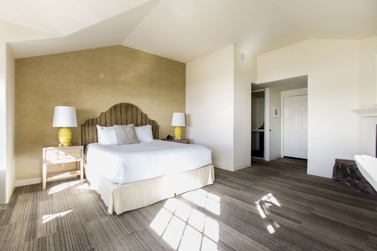 A spacious bedroom with a large bed, two bedside tables with lamps, a wooden headboard, and a view of a hallway.