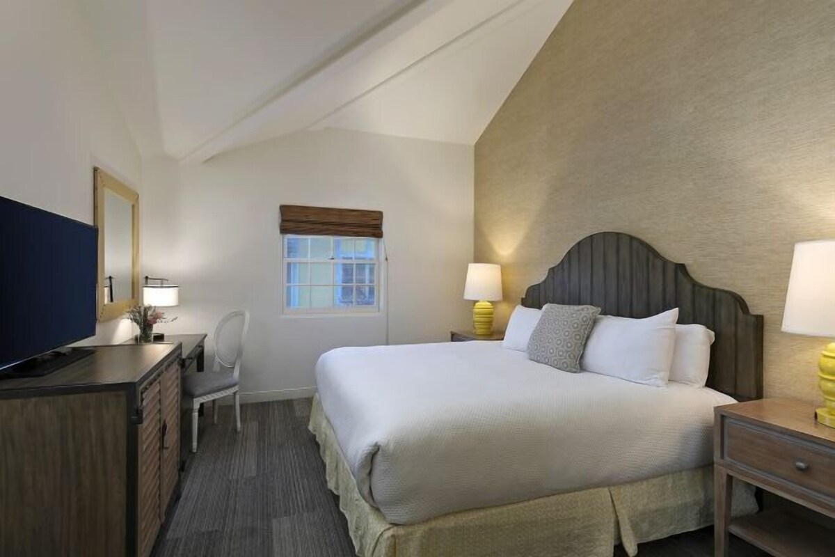 A hotel room with a bed, bedside tables, a desk, a chair, a television, and a window with blinds.