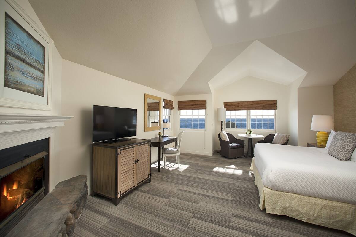 A hotel room with a fireplace, a television, a desk, a chair, a bed, and a view of the ocean through large windows.