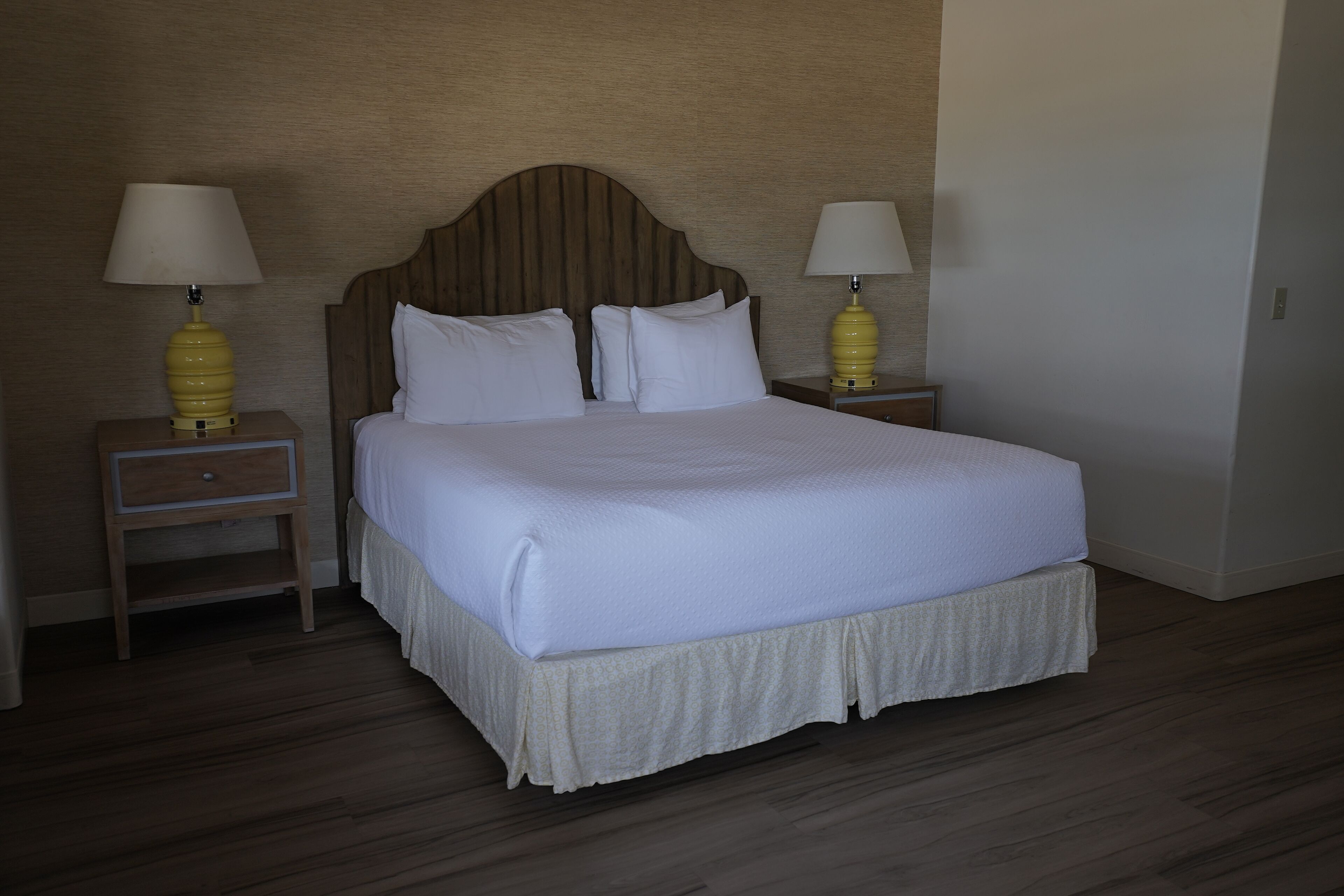 Una cama bien hecha con cabecera de madera, dos mesitas de noche con lámparas y suelo de madera.