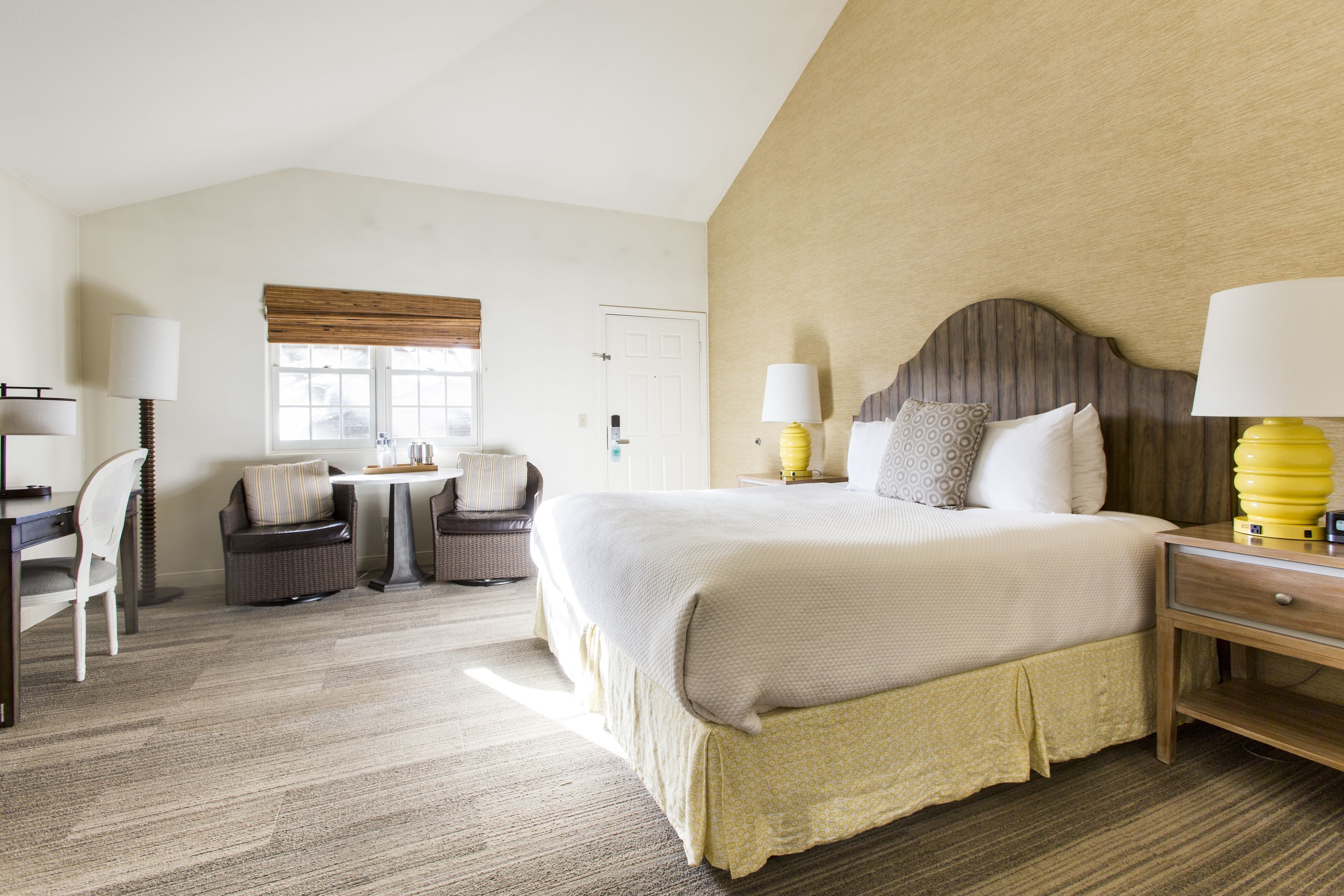 A hotel room with a large bed, a desk, two chairs, a window with blinds, and a lamp on the bedside table.