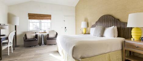 A hotel room with a large bed, a desk, two chairs, a window with blinds, and a lamp on the bedside table.