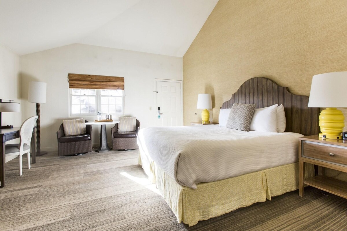 A hotel room with a large bed, a desk, two chairs, a window with blinds, and a nightstand with a lamp.