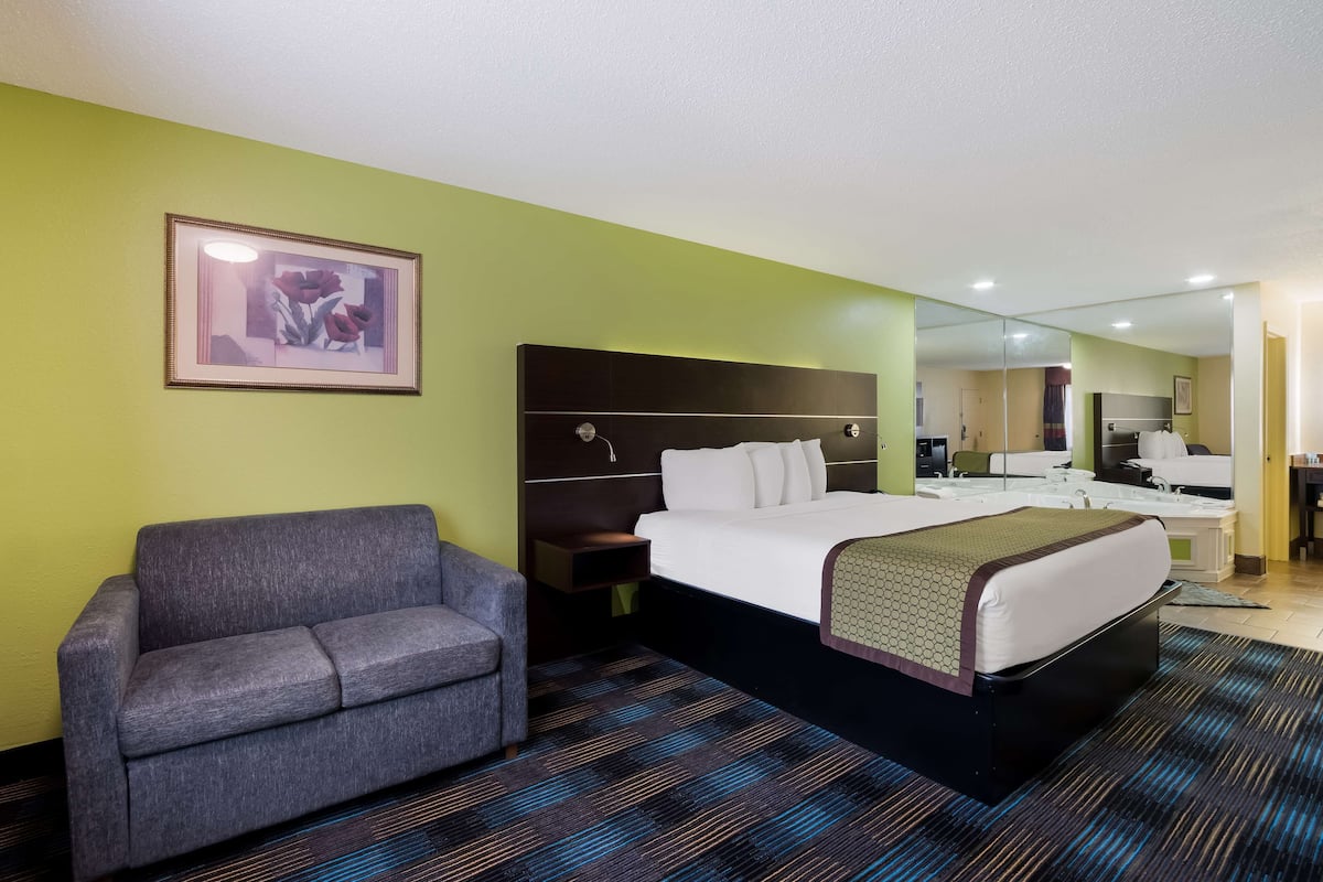 A hotel room with a bed, a sofa, a nightstand, a framed picture on the wall, and a bathroom visible through a mirror.
