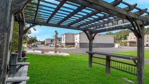 Una pérgola de madera con un banco, un césped cuidado y un estacionamiento con edificios al fondo.