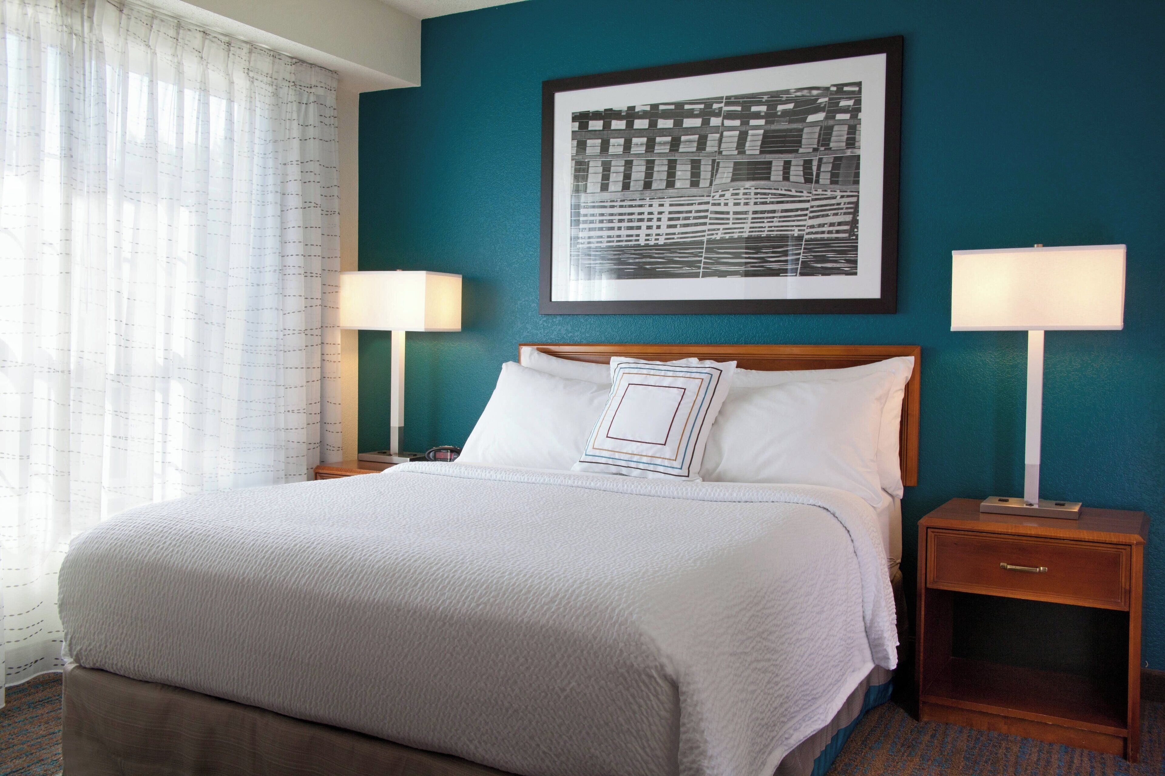 A neatly made bed with white linens, two bedside lamps, a framed picture on the wall, and a wooden nightstand with a drawer.