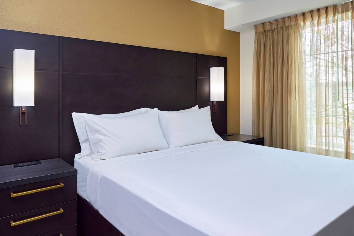 A hotel room with a large bed, dark headboard, bedside lamps, and a window with curtains.