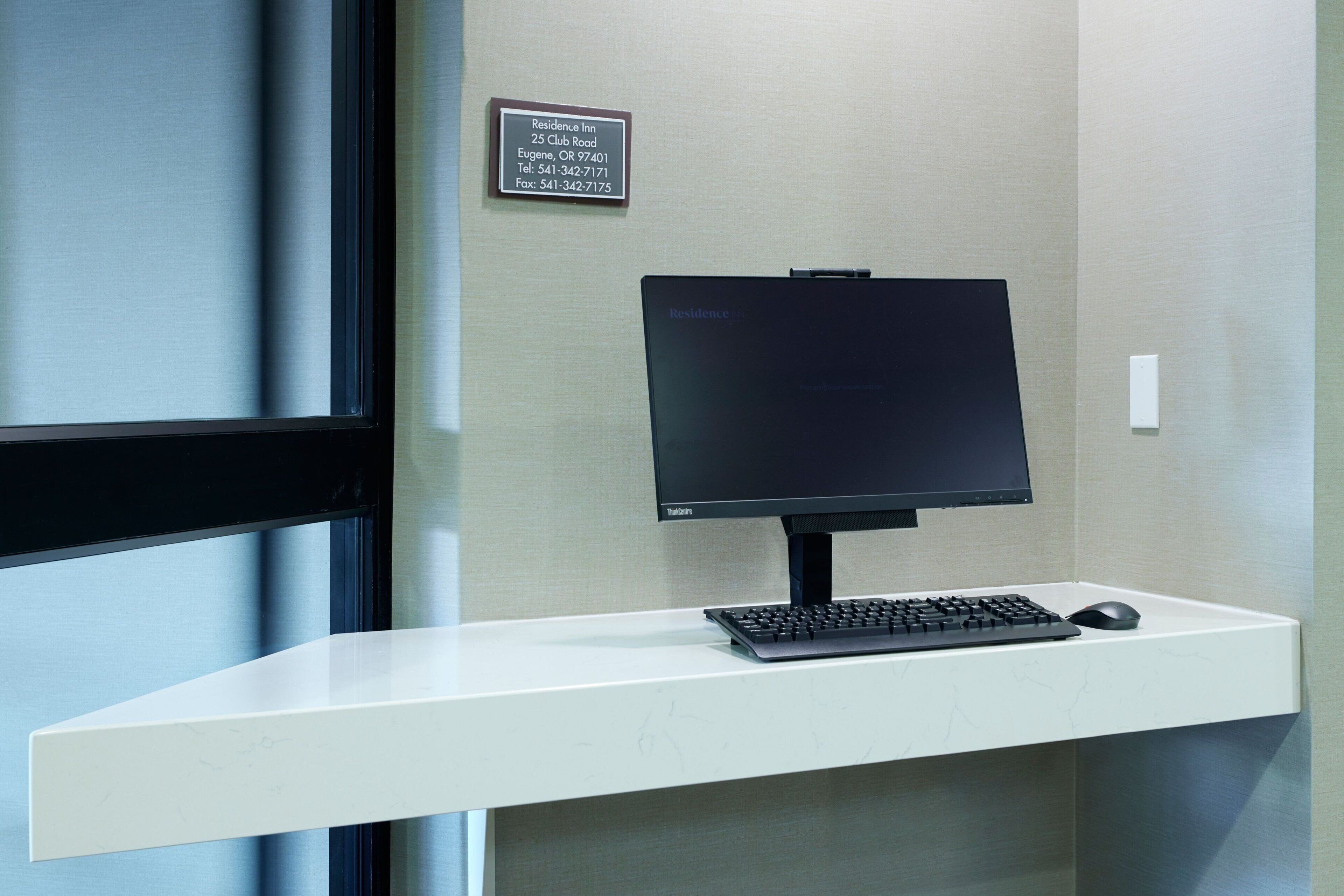 Ein moderner Schreibtisch mit Computerarbeitsplatz, inklusive Bildschirm, Tastatur und Maus. Ein Schild an der Wand gibt die Adresse und Kontaktinformationen des Residence Inn an.