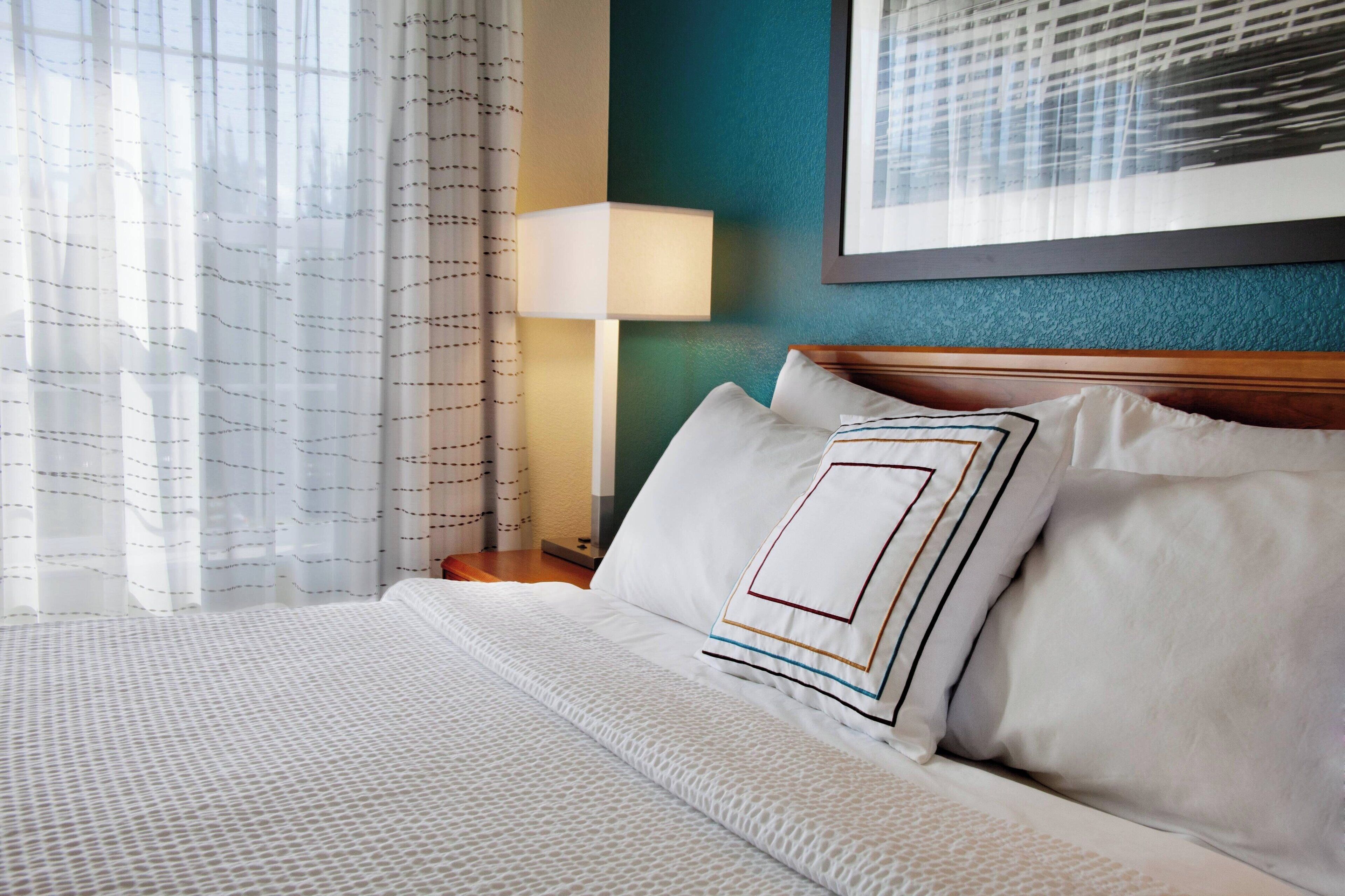 A hotel room with a neatly made bed, a nightstand with a lamp, and a framed picture on the wall.
