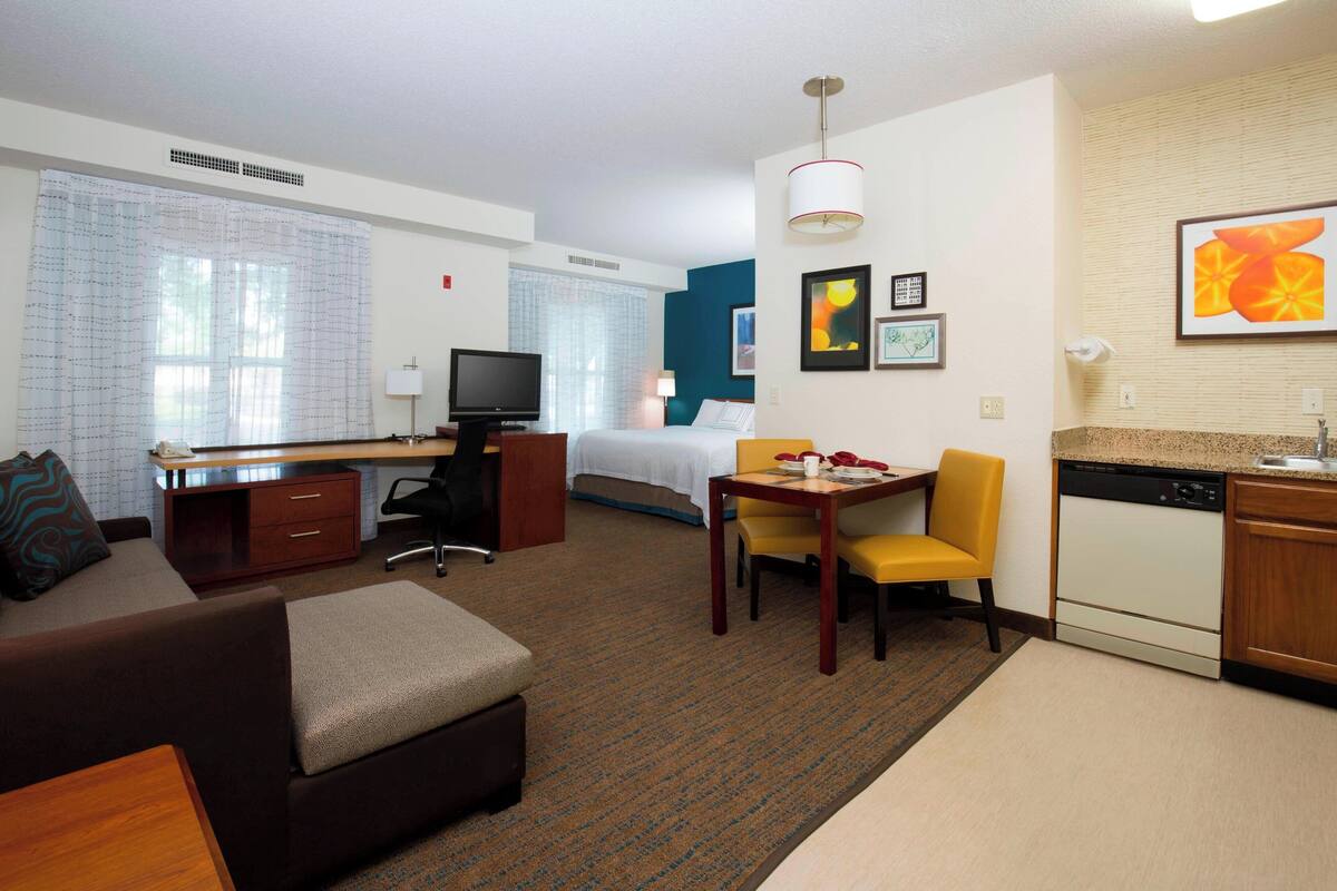 Une chambre d’hôtel comprenant un lit, un bureau avec un ordinateur, un canapé, une table à manger avec des chaises, un coin cuisine avec un lave-vaisselle et des œuvres d’art accrochées aux murs.