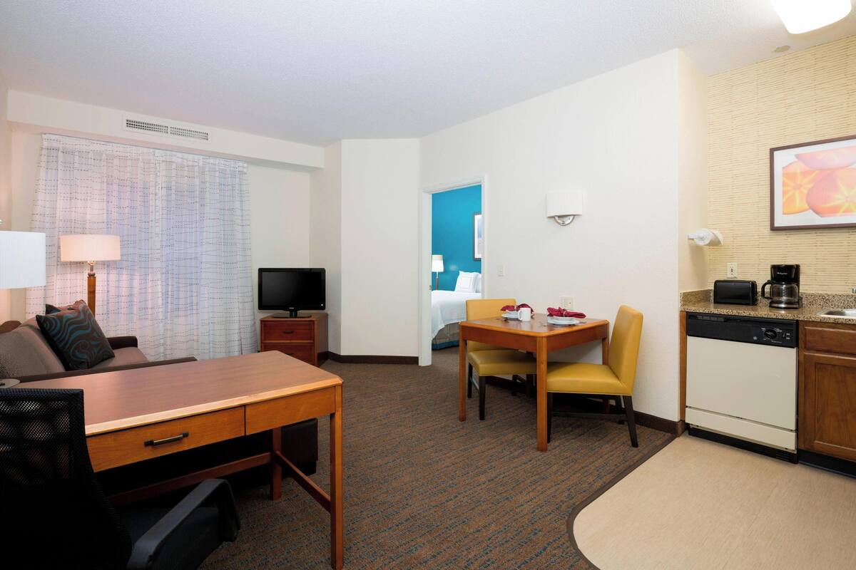 Une chambre d’hôtel avec un bureau en bois, une télévision, une table à manger avec des chaises, un coin cuisine avec une machine à café et un lit à l’arrière-plan.
