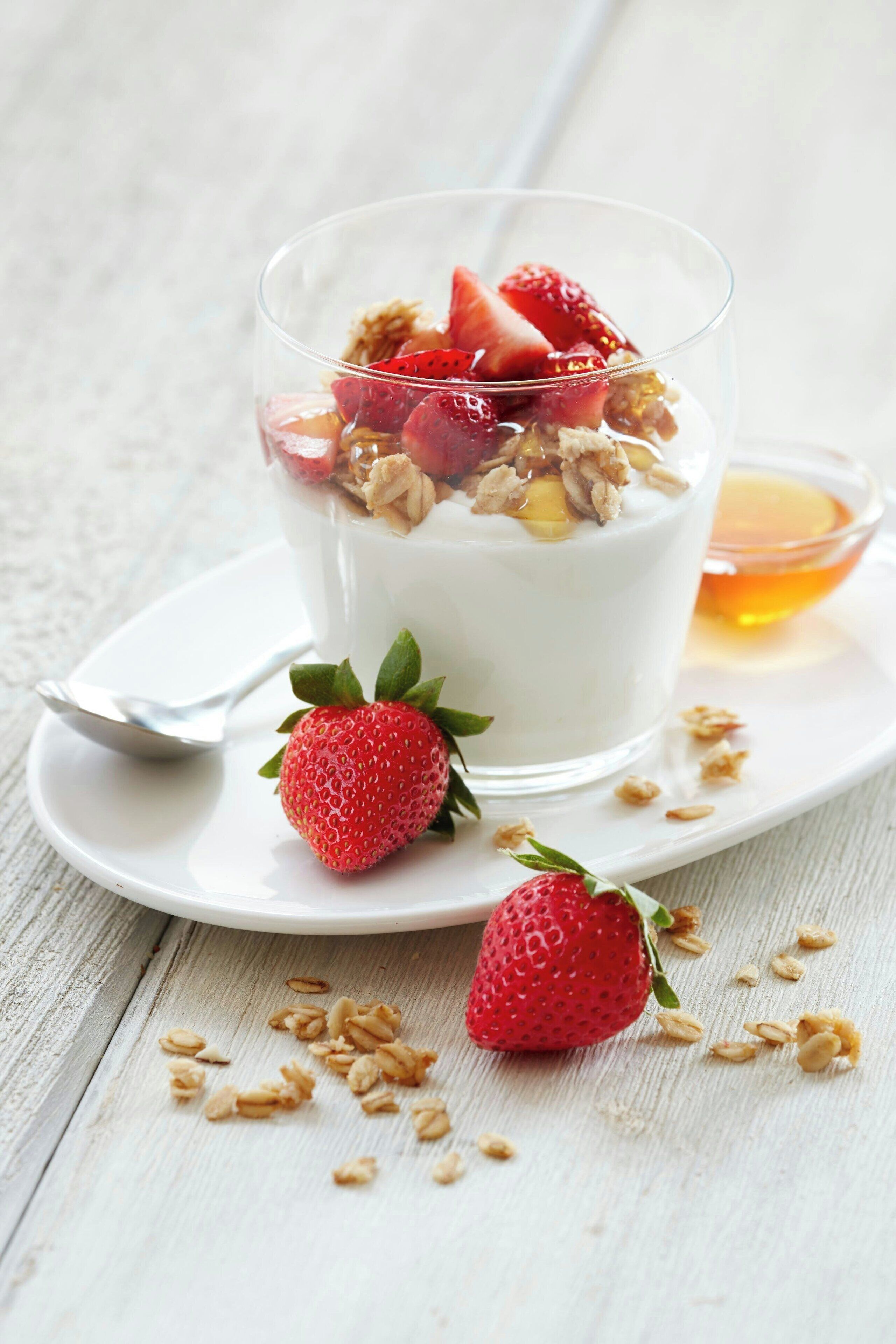 Un vaso de yogur coronado con granola y fresas, una fresa entera y un tazón pequeño de miel sobre un plato blanco.