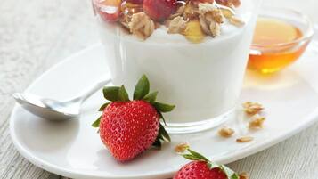 Un vaso de yogur coronado con granola y fresas, una fresa entera y un tazón pequeño de miel sobre un plato blanco.