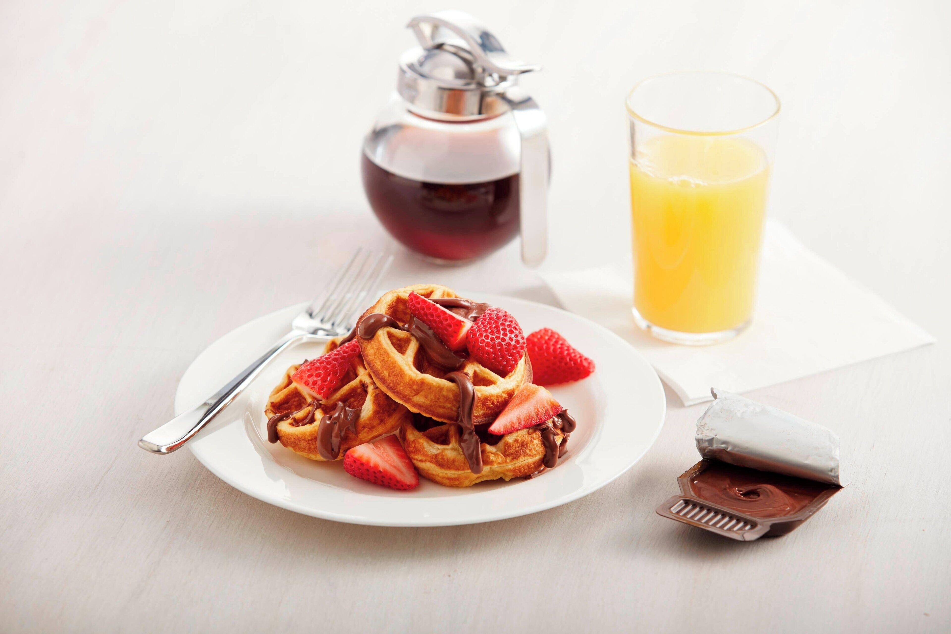 A plate of waffles with strawberries and chocolate sauce, a glass of orange juice, and a honey dispenser on a white table.