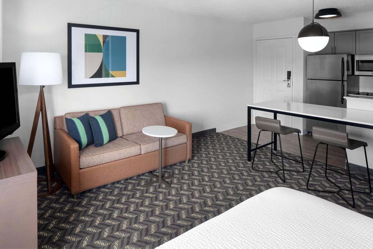 A modern hotel room with a sofa, a small round table, a TV, a kitchen area with a refrigerator and microwave, and a patterned carpet.