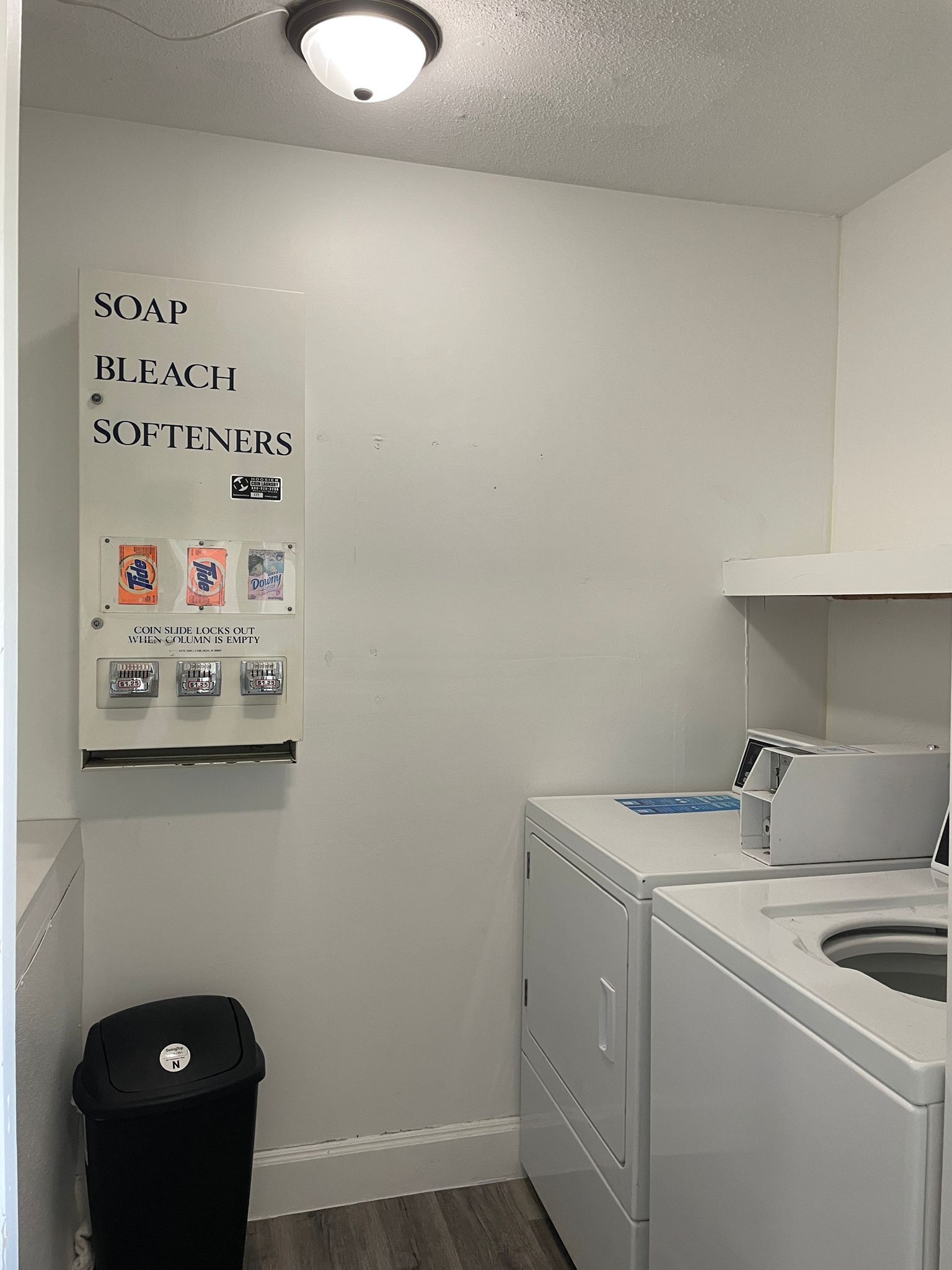 A laundry room with a wall-mounted soap and bleach dispenser, a coin-operated dryer, and a trash bin.