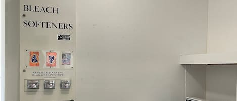 A laundry room with a wall-mounted soap and bleach dispenser, a coin-operated dryer, and a trash bin.
