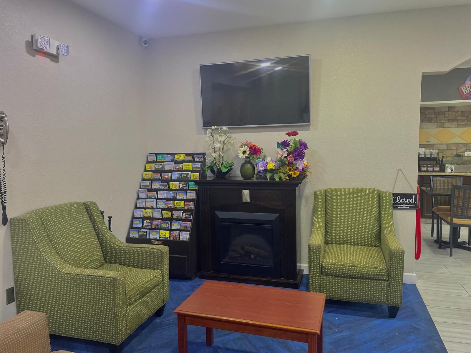 Una habitación con dos sillones verdes con estampado, una mesa de centro de madera, una chimenea con un televisor encima, un arreglo floral y un cartel de cerrado en la pared.