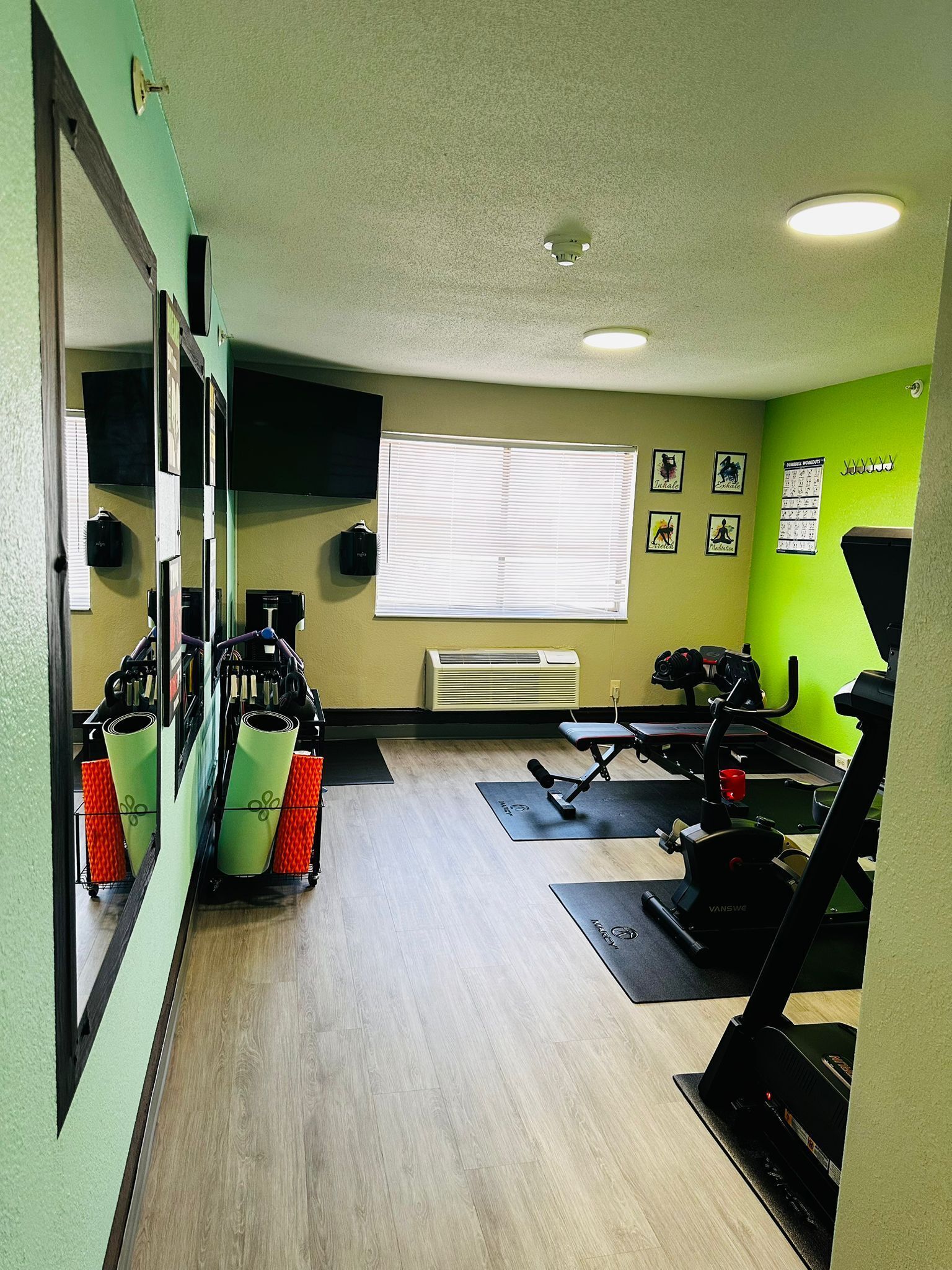 Un gimnasio con equipo de ejercicio, un ventanal con persianas y un espejo en la pared.