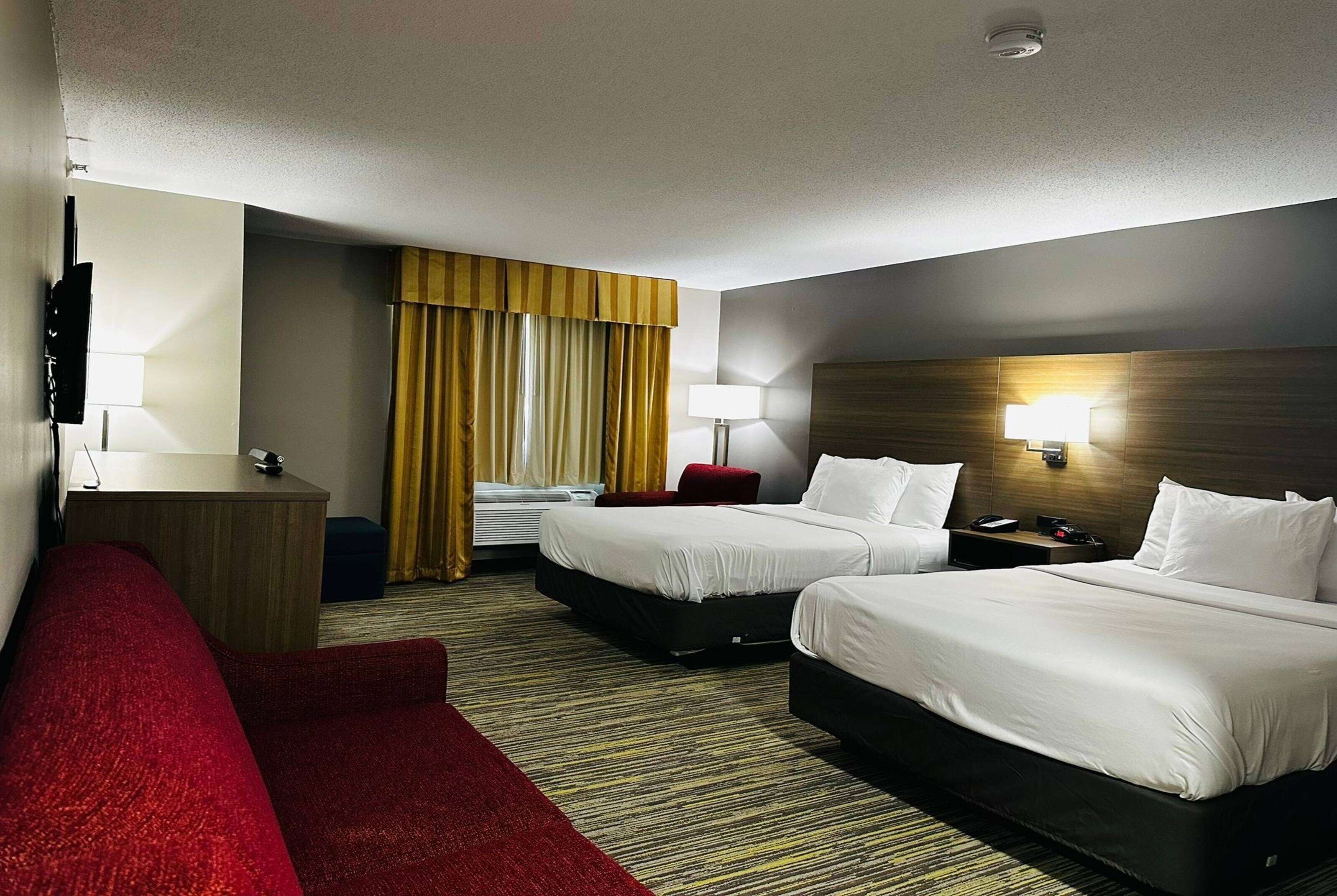 Une chambre d’hôtel avec deux lits, un bureau, un canapé rouge, une lampe et une fenêtre avec des rideaux.