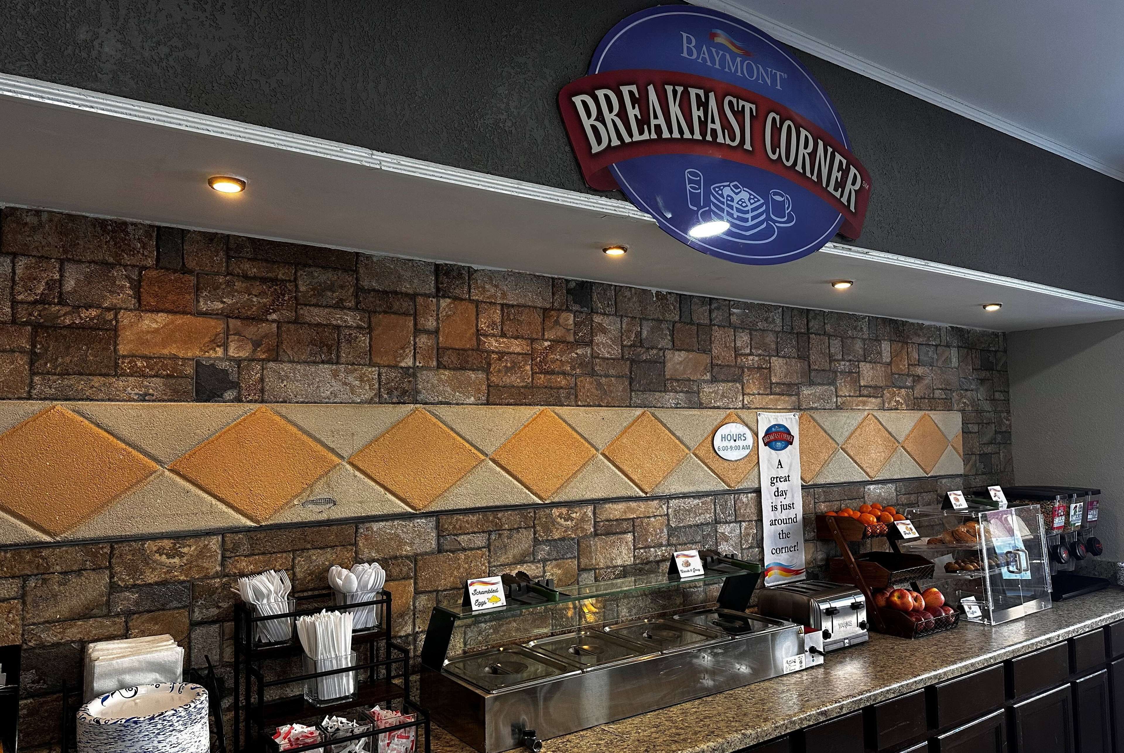 A breakfast counter with a sign reading "Baymont Breakfast Corner," a stone wall with a diamond pattern, and various breakfast items displayed.