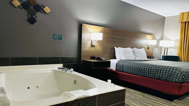A hotel room with a large bathtub, a bed with a red and black patterned bedspread, two bedside lamps, and a wooden headboard. - Baymont by Wyndham Anderson (Anderson)