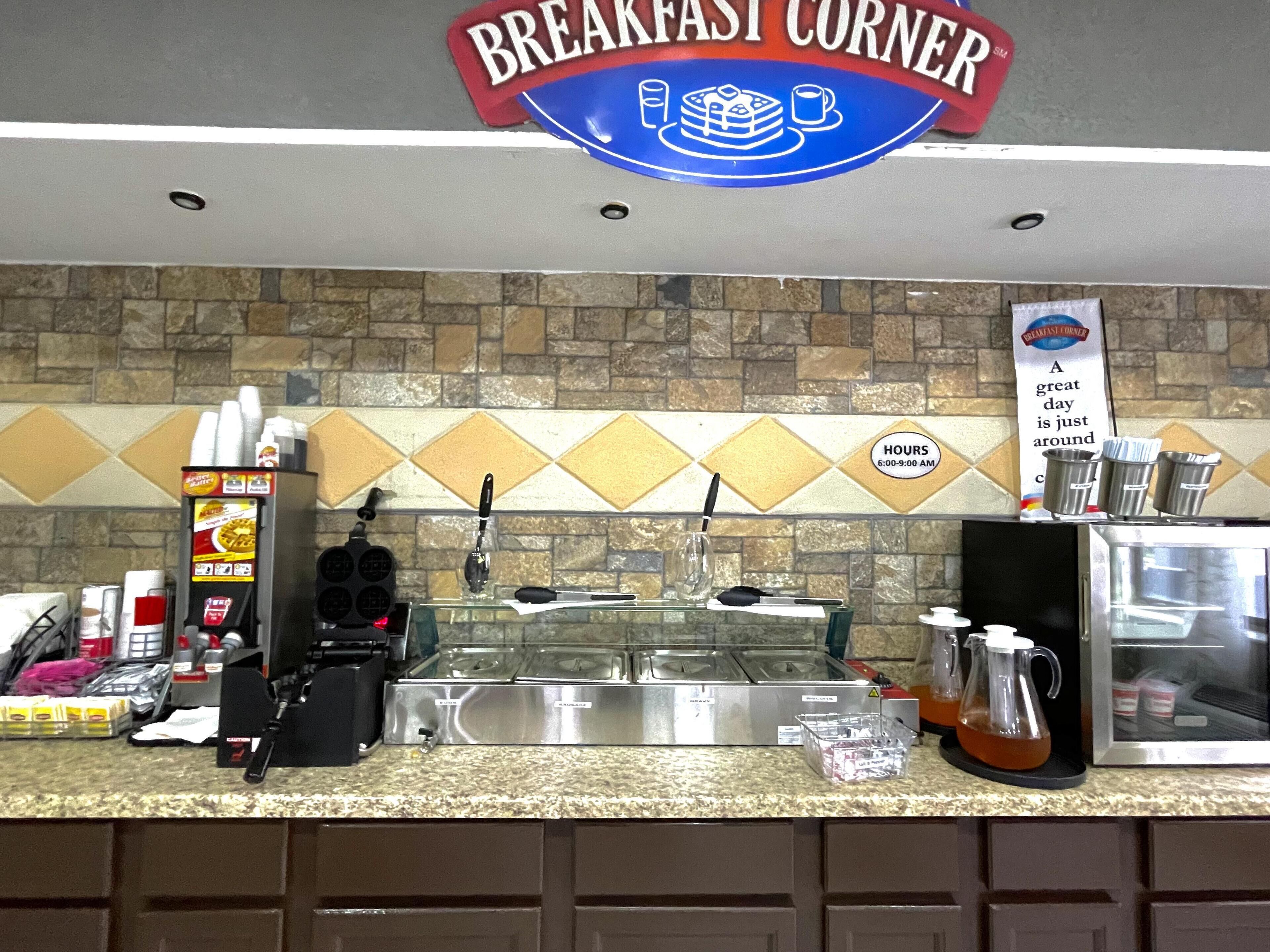 Un coin petit-déjeuner avec un panneau indicateur, une machine à café, une bouilloire et un tableau affichant le menu.