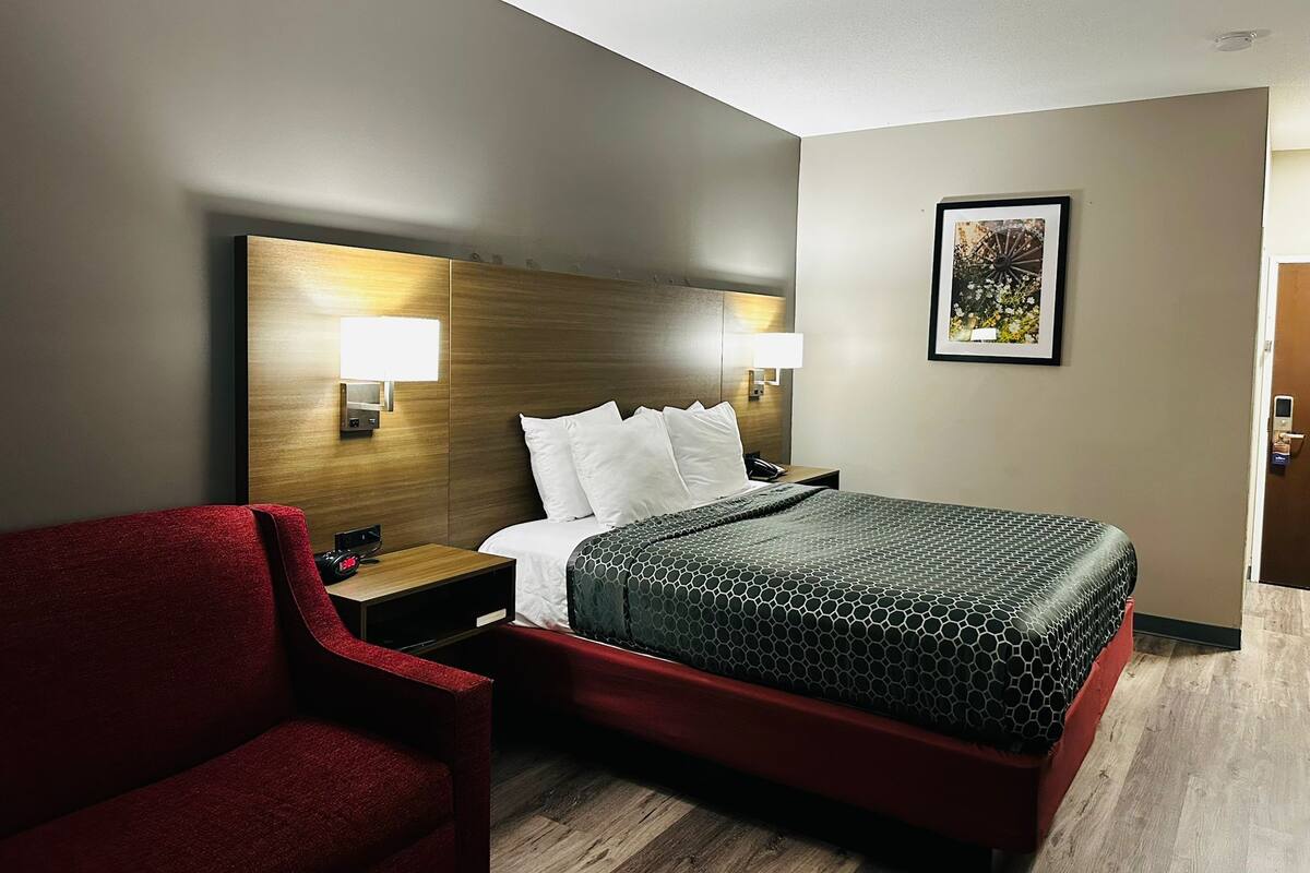 Habitación de hotel con cama, un sillón rojo, cabecera de madera, mesita de noche, lámpara y un cuadro en la pared.