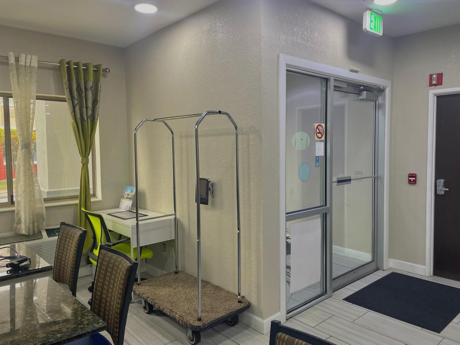 A hotel room with a dining area, a cart, a glass door, and an exit sign.