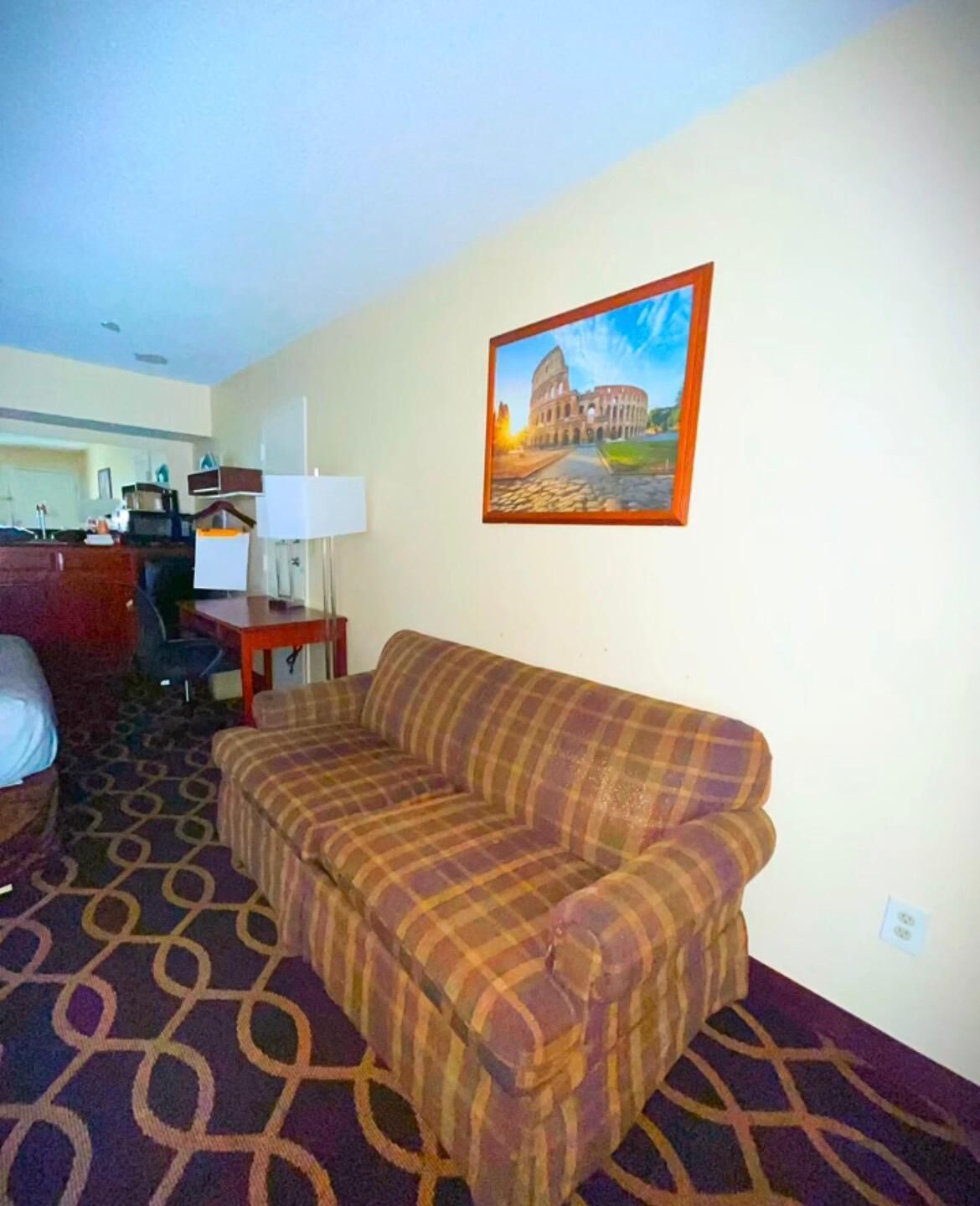 A hotel room with a plaid sofa, a desk with a lamp, and a painting of the Colosseum.
