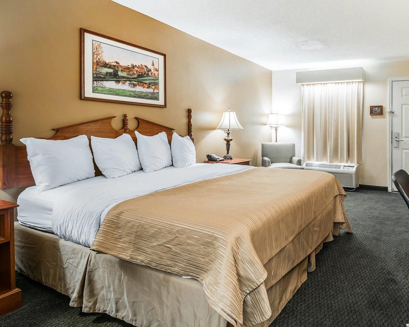 A hotel room with a large bed, a bedside table with a lamp, a chair, a window with curtains, and a painting on the wall.